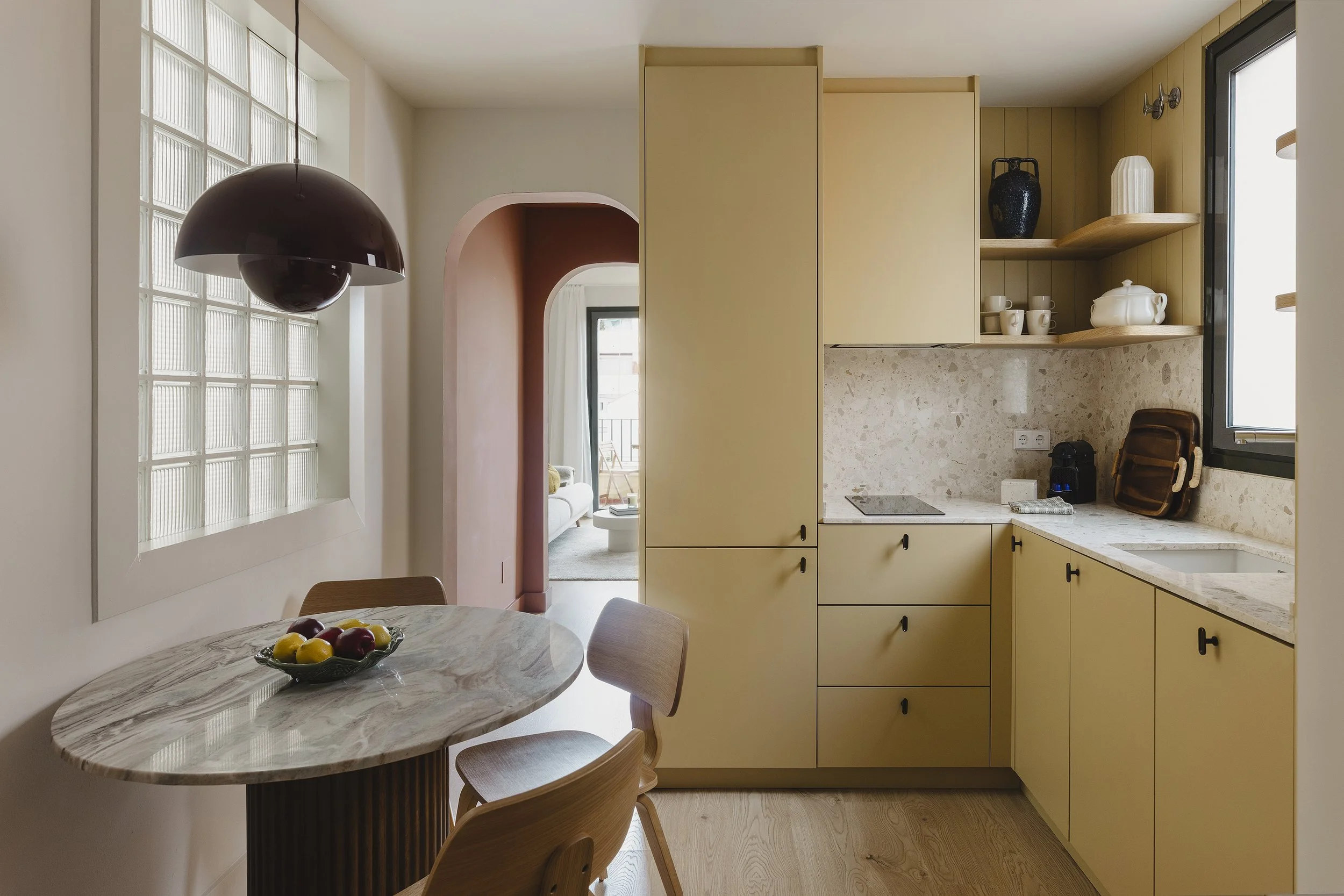 Cocina con armarios amarillos, encimera de mármol, estanterías abiertas con platos y un jarrón negro, ventana y una mesa de comedor con un frutero y sillas.