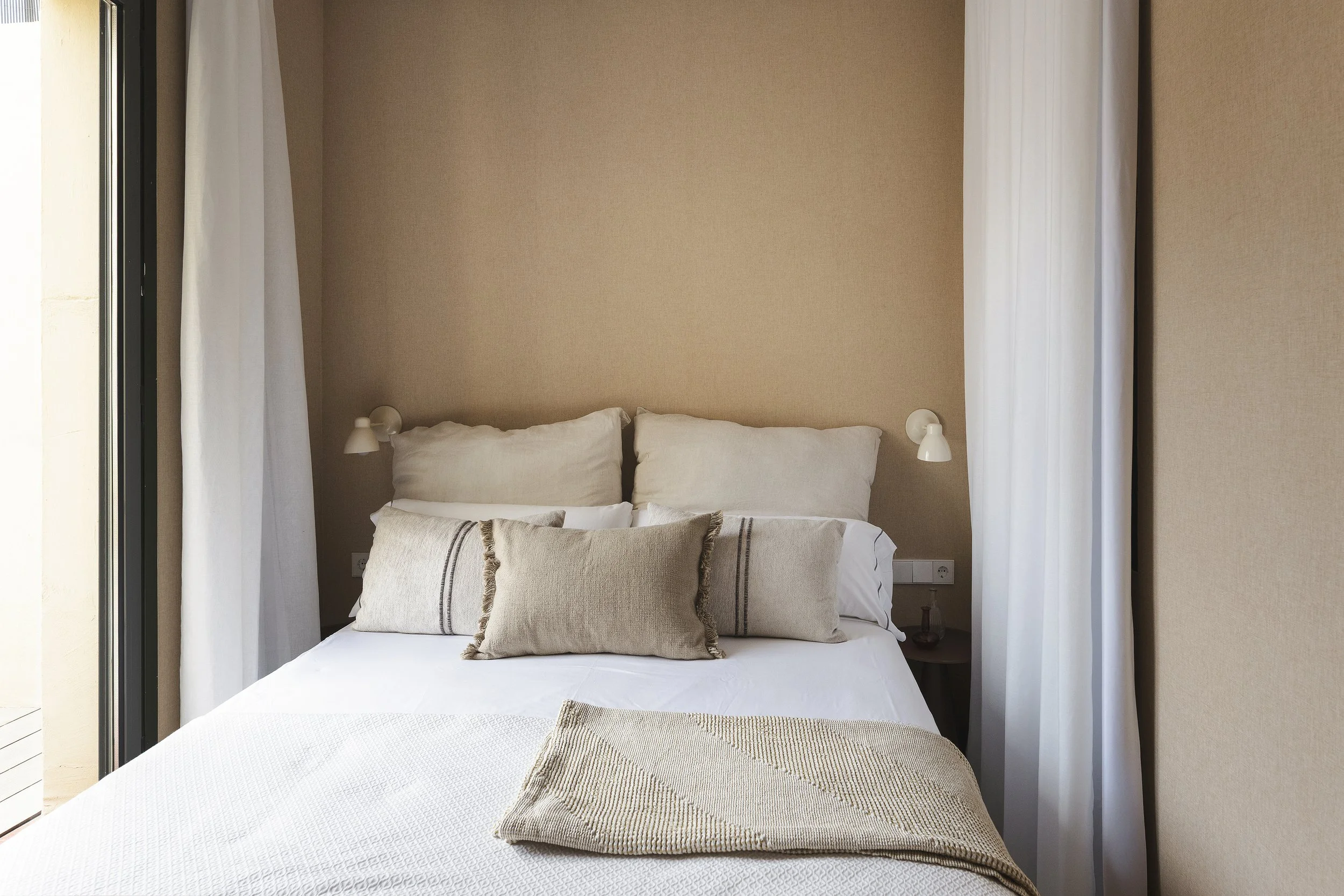Una cama cuidadosamente confeccionada con ropa de cama blanca, dos grandes almohadas blancas y tres cojines decorativos beige con detalles de flecos. La cama está situada contra una pared beige con dos lámparas de lectura montadas en la pared a cada lado. Cortinas de color claro cubren la cama.