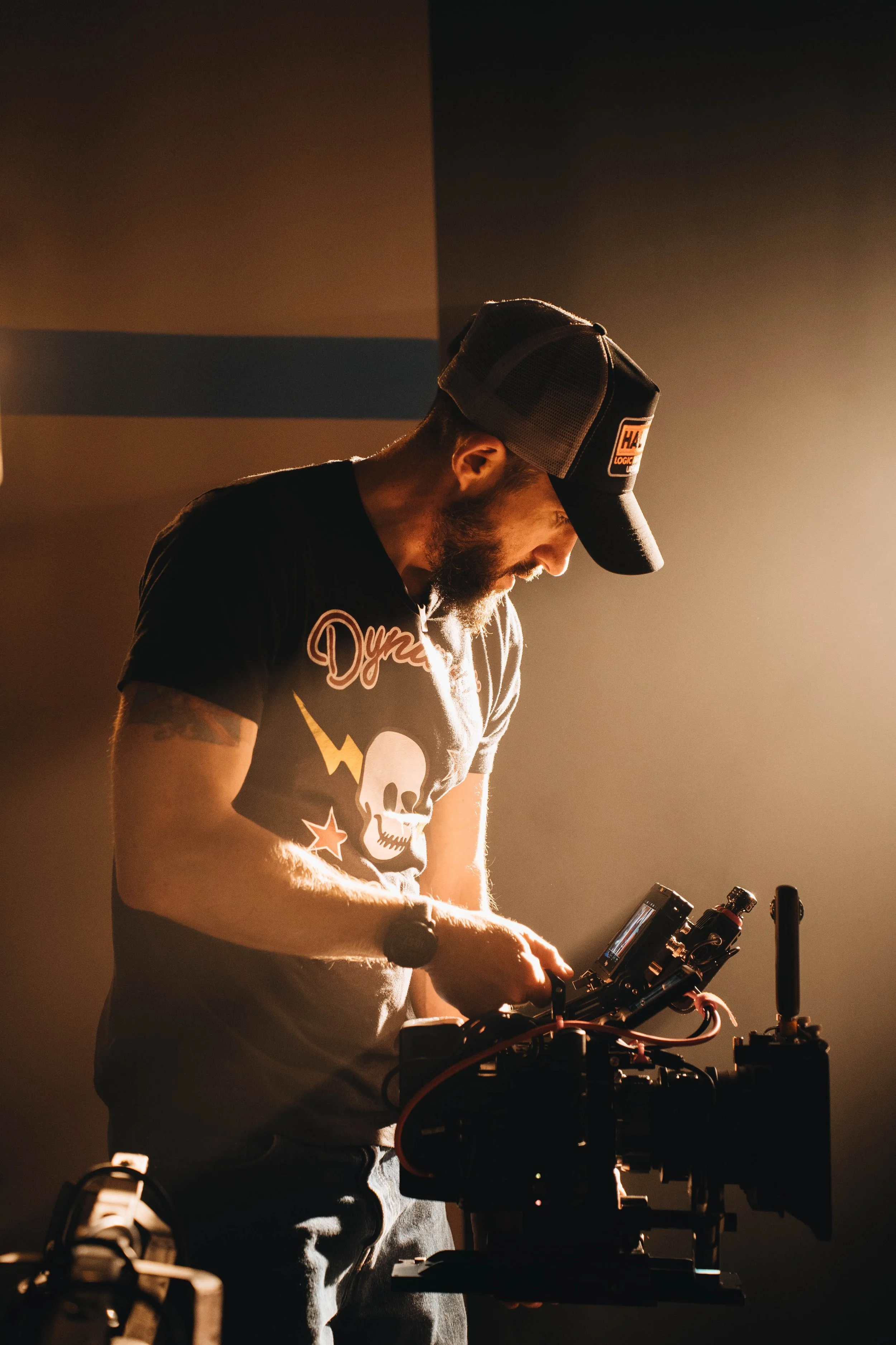A man working on a professional camera setup, wearing a black T-shirt with a skull, lightning bolt, star graphics, and a black cap, illuminated by warm lighting.