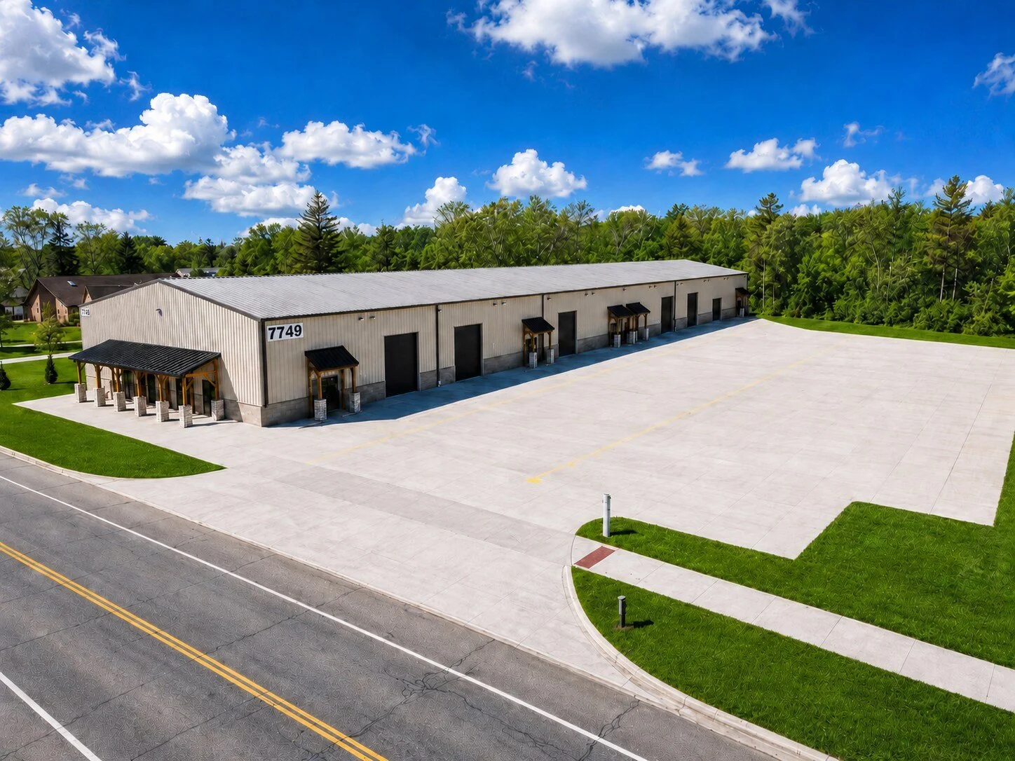 A large, empty industrial warehouse with multiple loading docks, situated in a spacious, paved lot near a street, surrounded by green grass and trees, under a bright blue sky with scattered clouds.