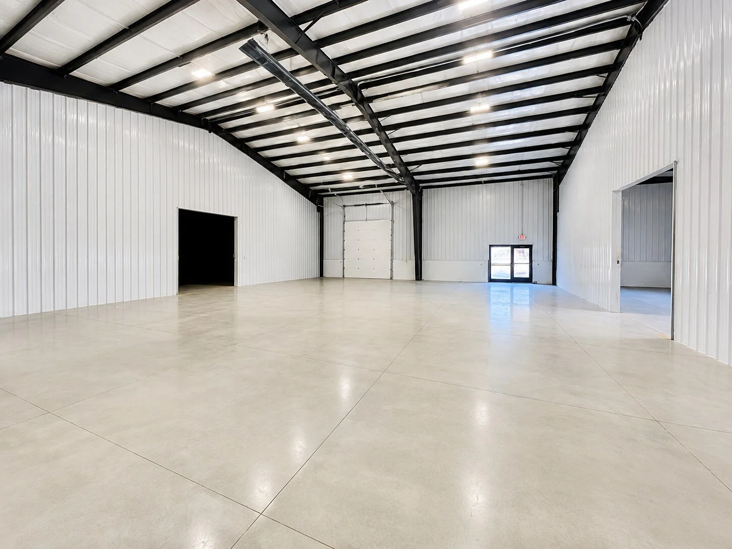 Empty industrial warehouse with polished concrete floors, white metal walls, black steel beams, and a high ceiling with lighting. Doors include a large roll-up and a glass entrance door.