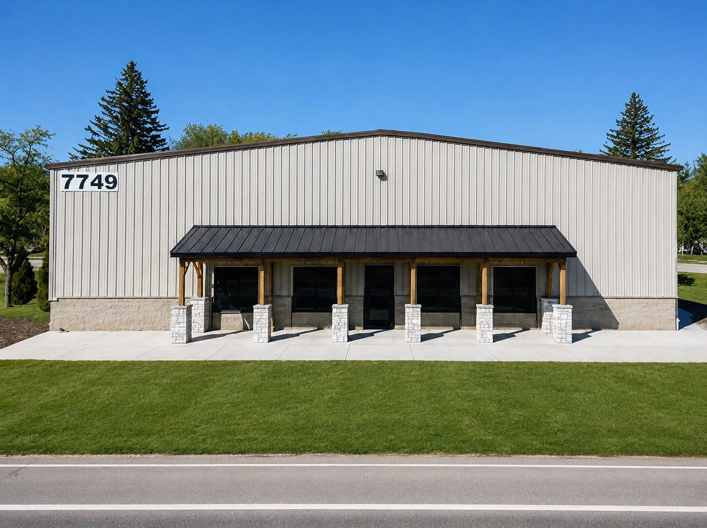 Commercial building with white metal exterior and four large windows shaded by a black metal roof, located at 7749, along a sidewalk and road, with green lawn and trees in the background.