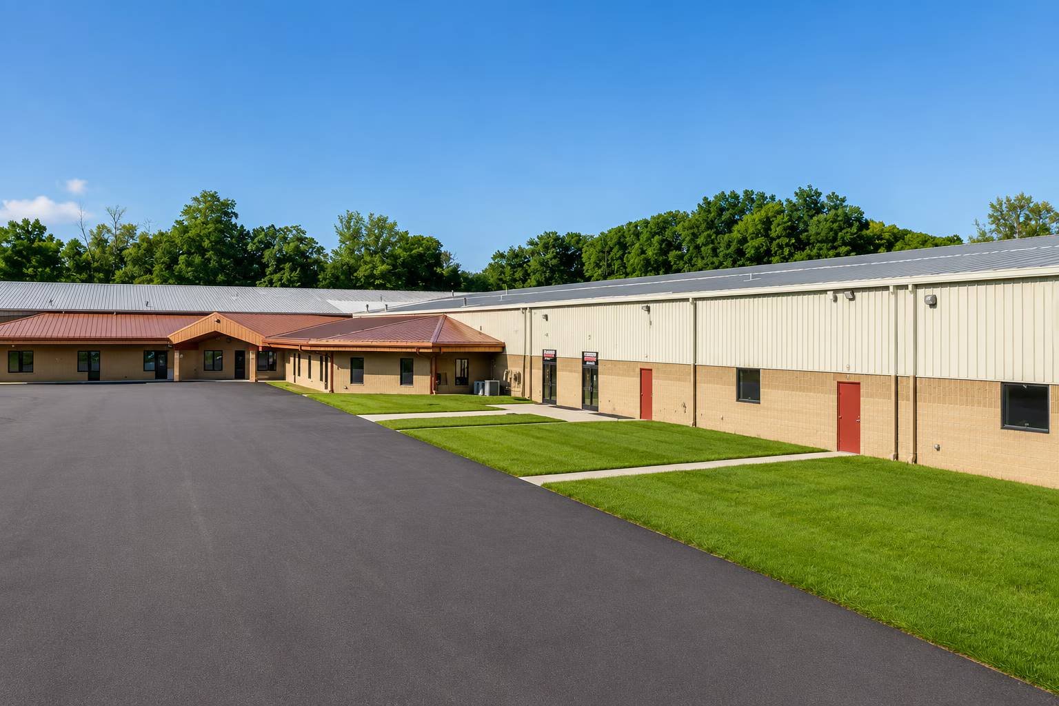 A large building with a beige and brick exterior, metal roof, and red doors, surrounded by a parking lot and green grass, with trees and a blue sky overhead.