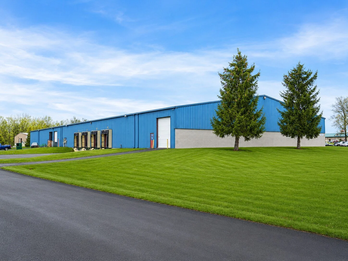 Blue industrial warehouse building with green lawn and trees in the foreground under a blue sky with scattered clouds.