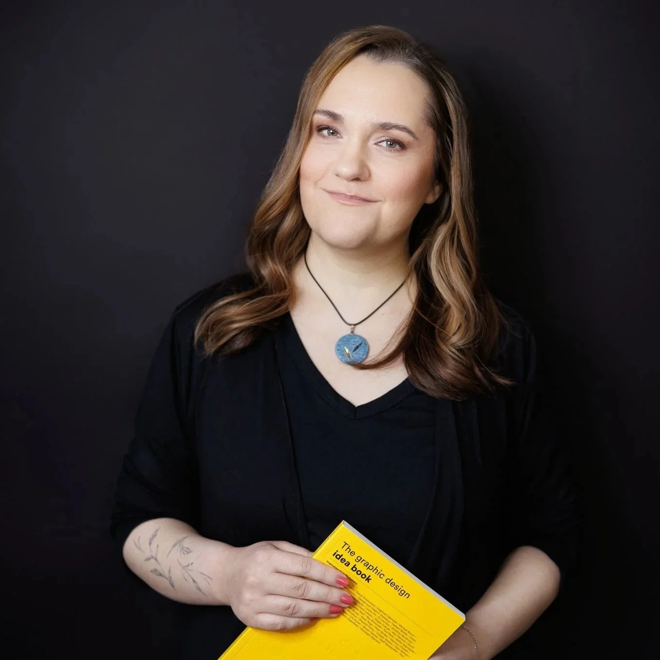 A woman with brown, wavy hair wearing a black top and a circular blue necklace, holding a yellow book titled 'The graphic design idea book,' standing against a black background.
