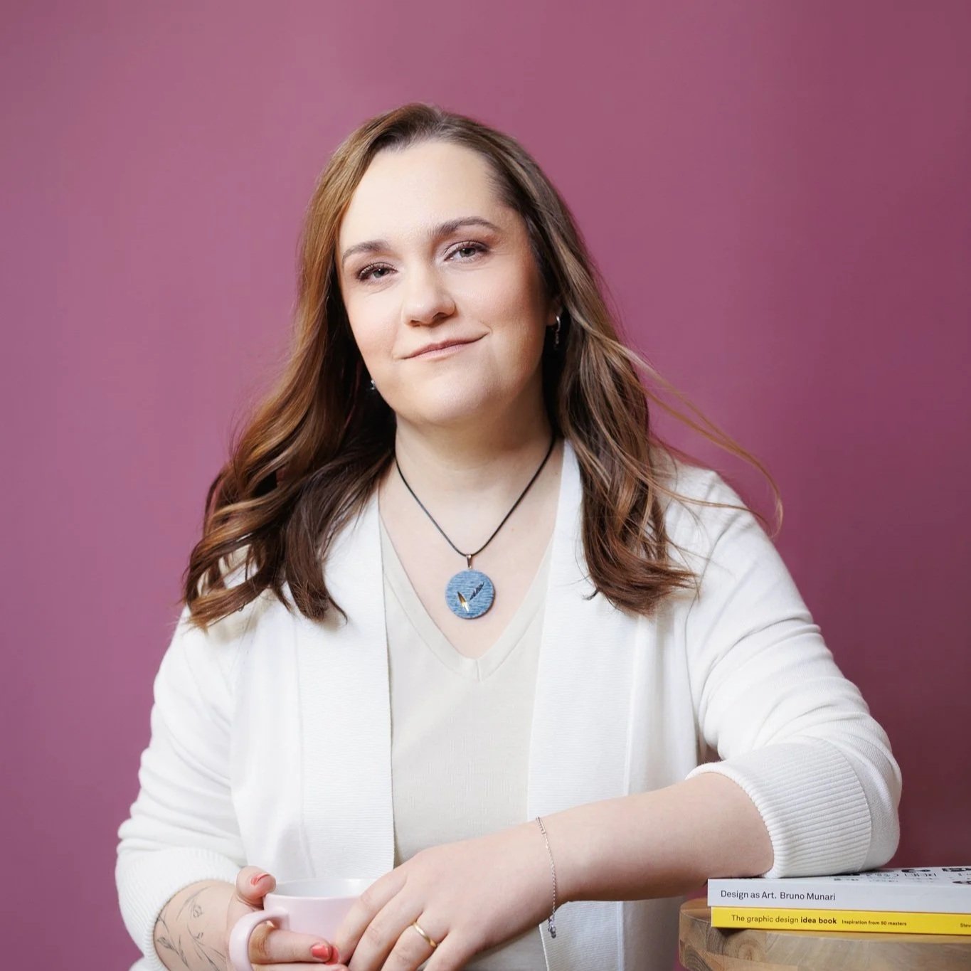 A woman with wavy brown hair and light skin, wearing a white shirt and cardigan, holds a pink mug, against a purple background, wearing a blue pendant necklace.