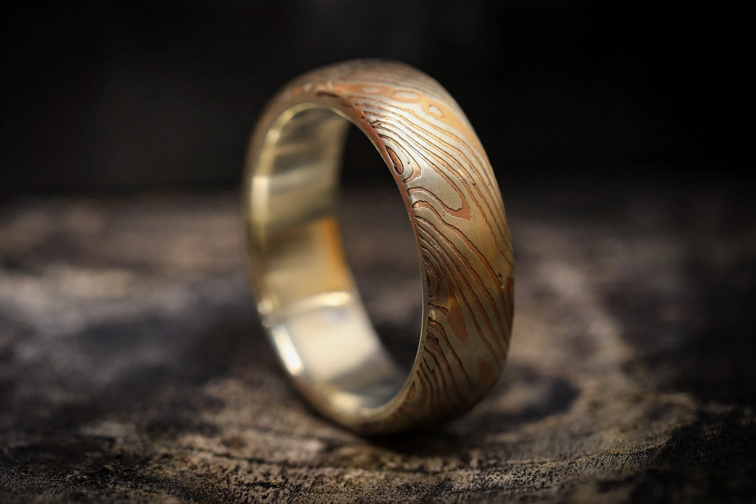 Close-up of a gold wedding band with a textured, wavy pattern resting on a dark, textured surface.