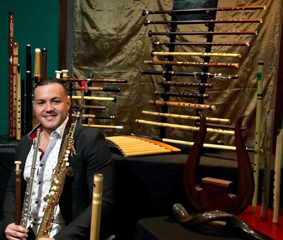 Man in black suit holding a gold-colored saxophone, standing in front of a display of various wooden flutes and musical instruments, with a gold curtain backdrop.