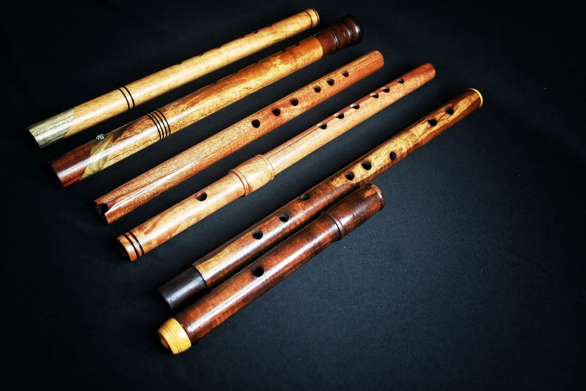 A collection of wooden handmade wooden ethnic wind instruments with varying lengths and wood finishes, laid out horizontally on a black surface.