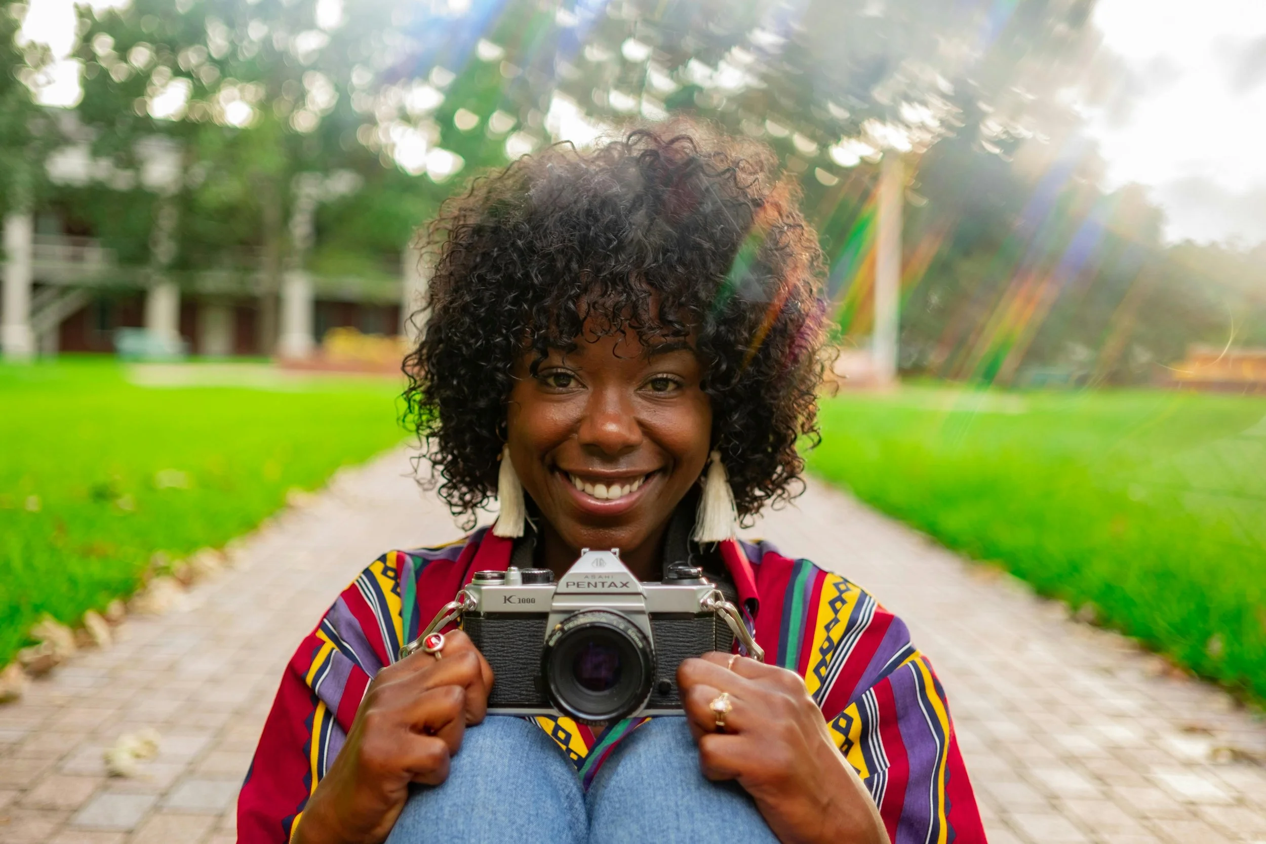 bipoc woman holding camera, sitting on ground, smiling