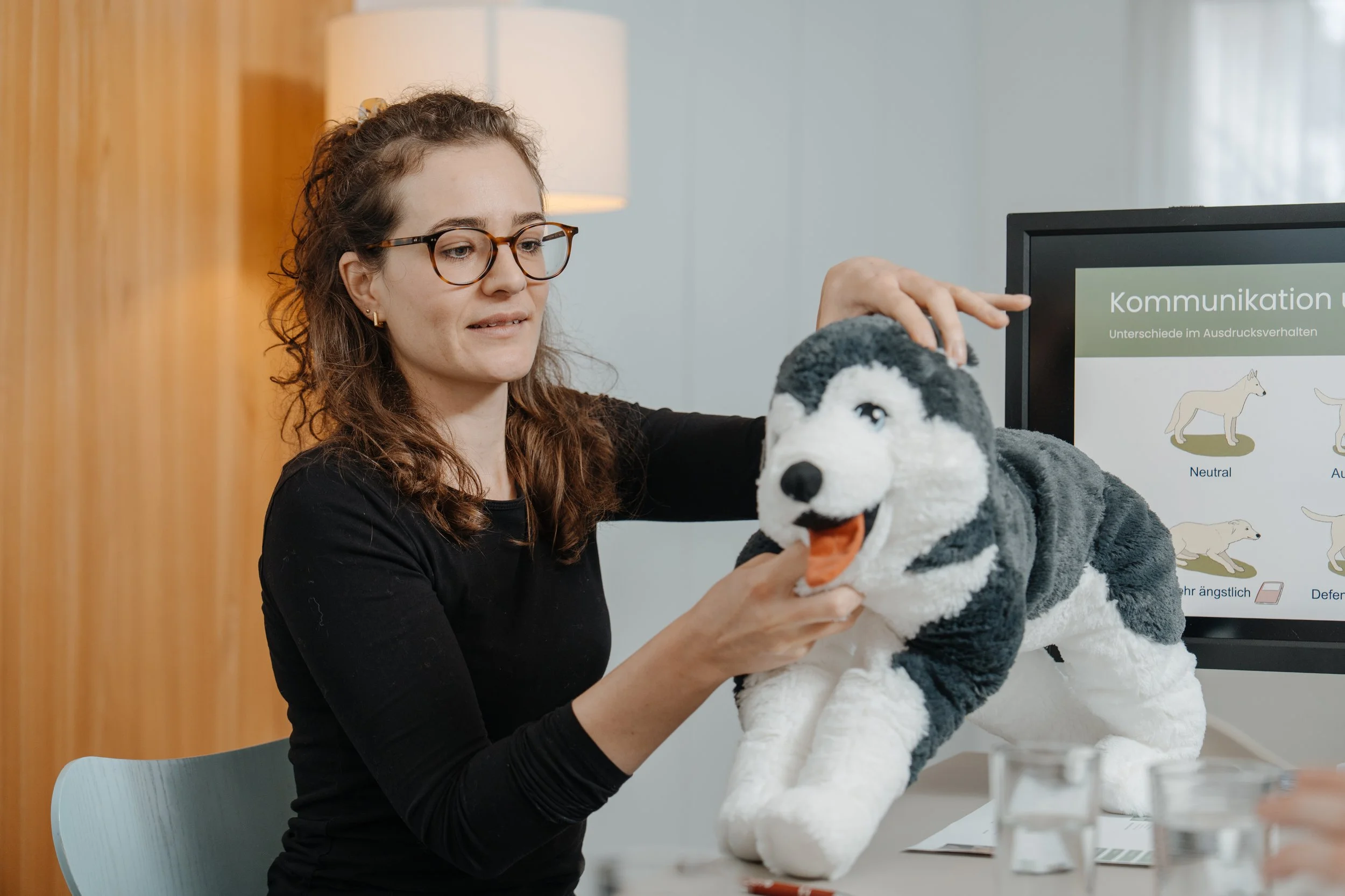 Talina Caviezel hält eine plüschigen Husky und erklärt die Körpersprache, während ein Präsentationsbildschirm im Hintergrund eine Folie zum Thema Kommunikation zeigt.