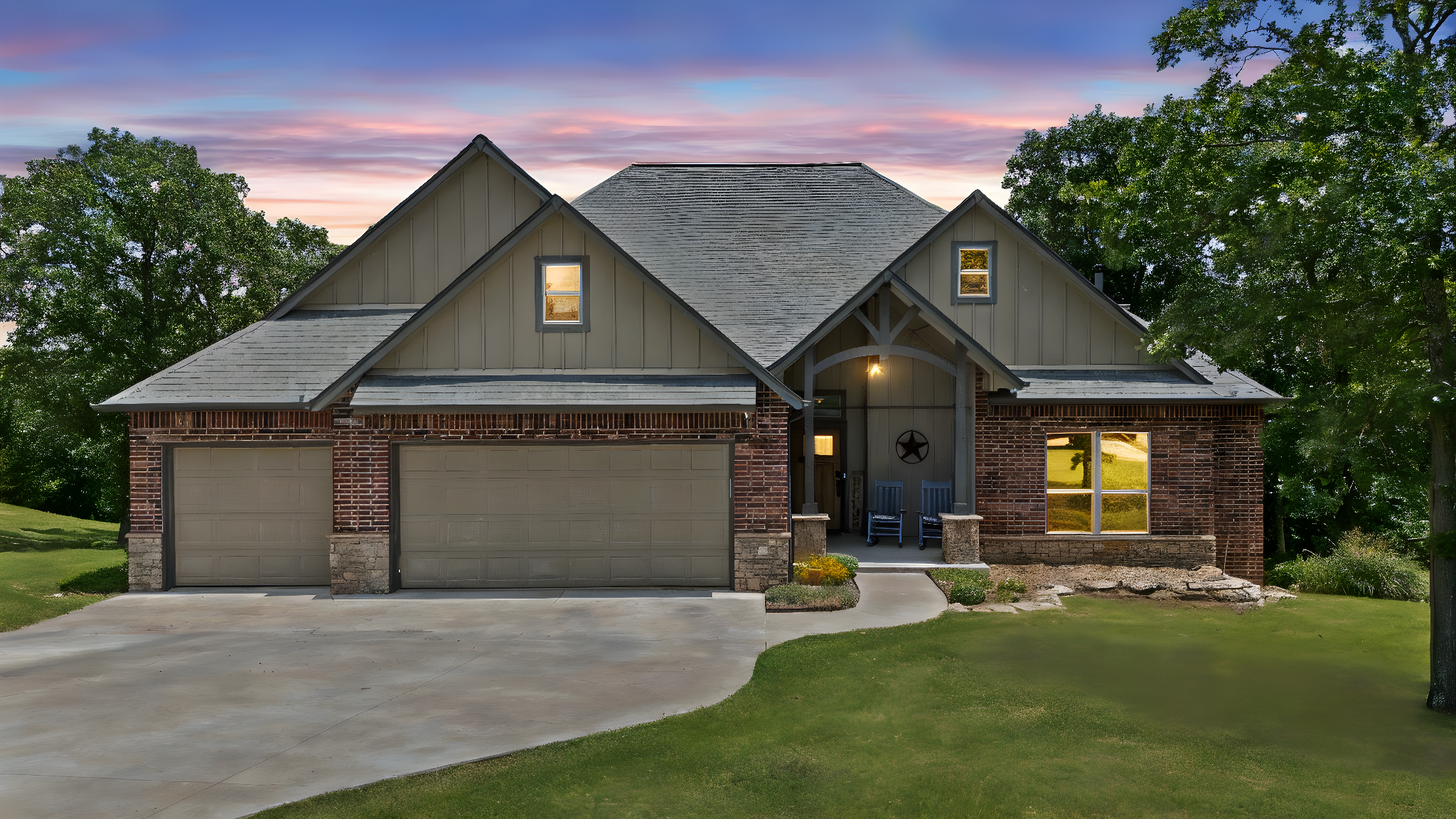 A two-story house with brick and siding exterior, a double garage, front porch with two chairs, surrounded by green lawns and trees, under a colorful sunset sky.