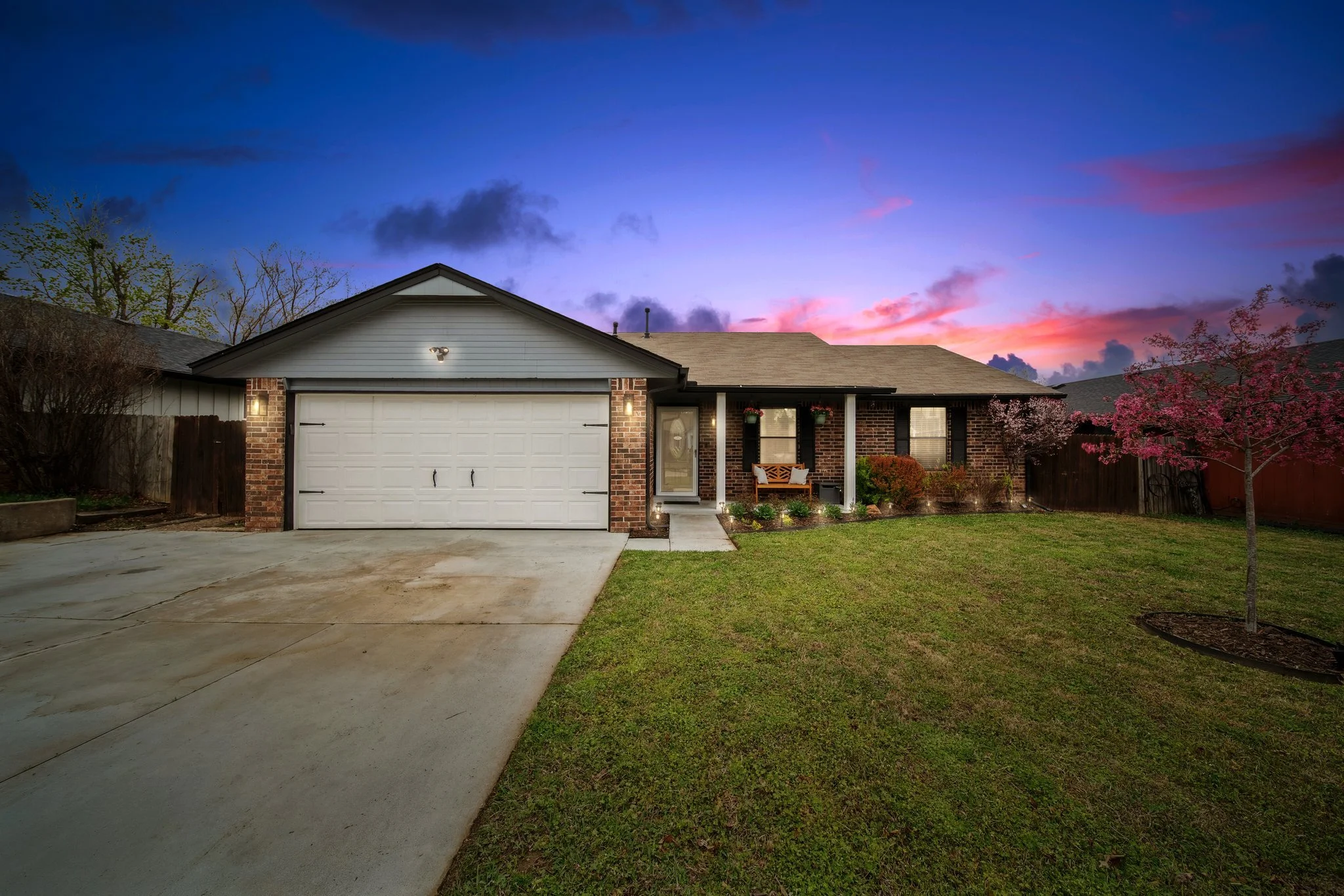 Front view of a house during twilight with a well-manicured lawn, flowering trees, and front porch lighting.