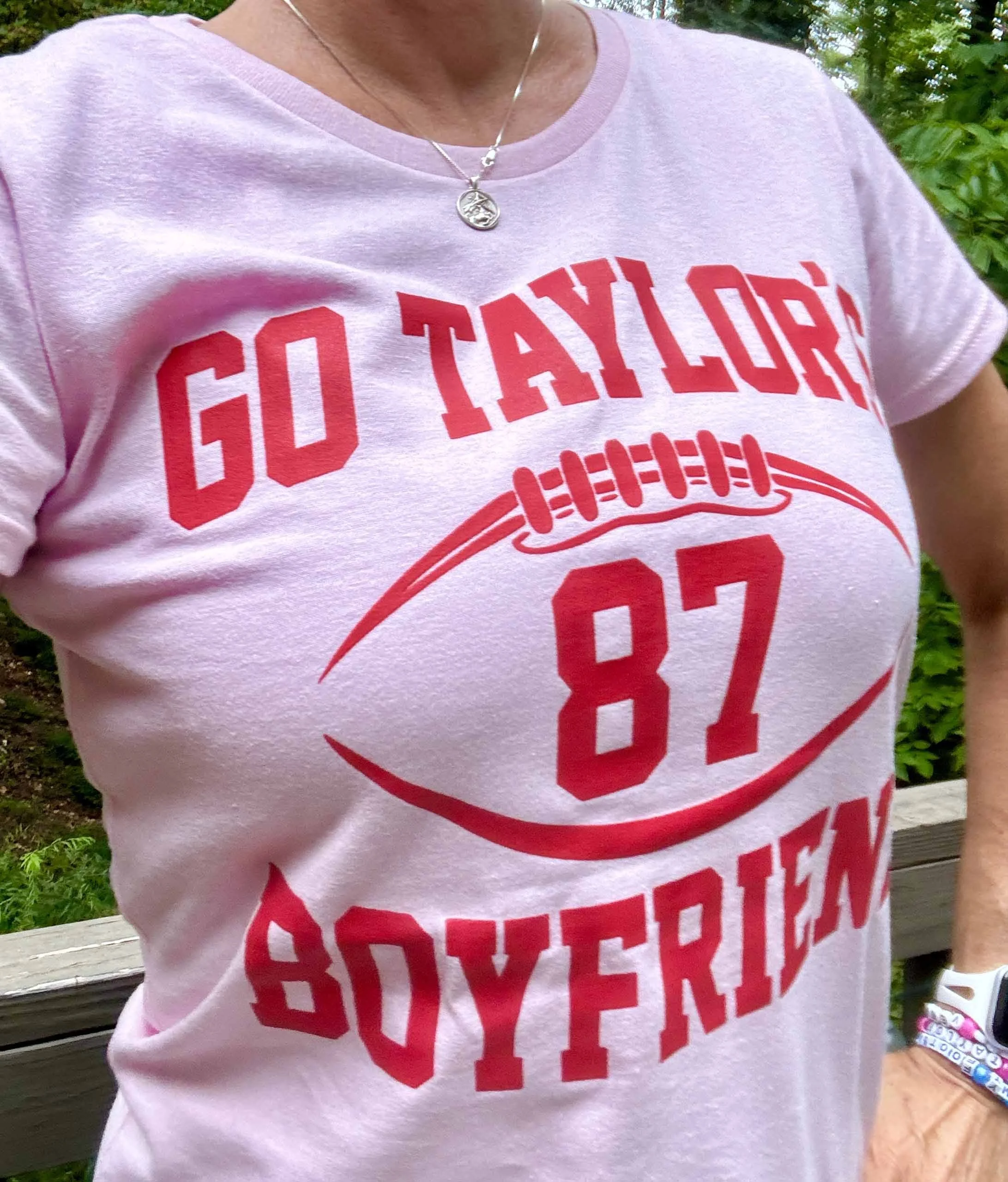 Close-up of a person wearing a pink T-shirt with red text reading 'Go Taylor, 87, Soyeon' and a small necklace with a circular pendant, outdoors with green foliage in the background.