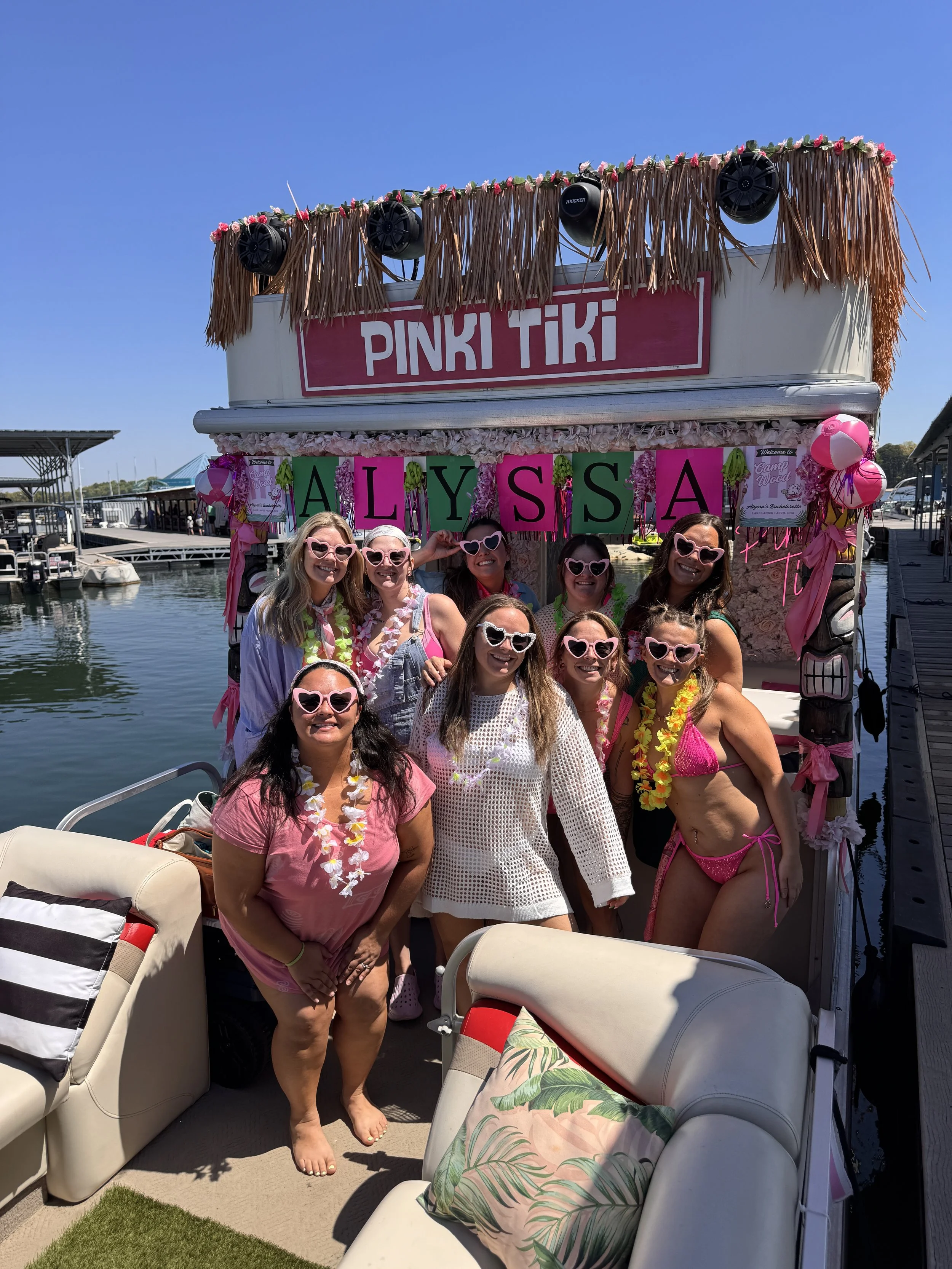 A group of women in pink swimsuits and summer attire enjoying a day at the beach, posing together in front of trees and sand, some with sunglasses, hats, and leis. One woman is lying on the sand with her legs raised, wearing a hat that says 'Bride'.