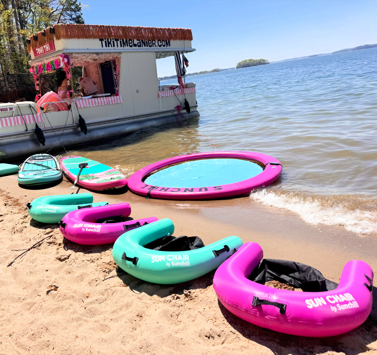 A floating tiki bar on a lake with pink and green inflatable sun chairs and a pink inflatable pool ring on the sandy shore.
