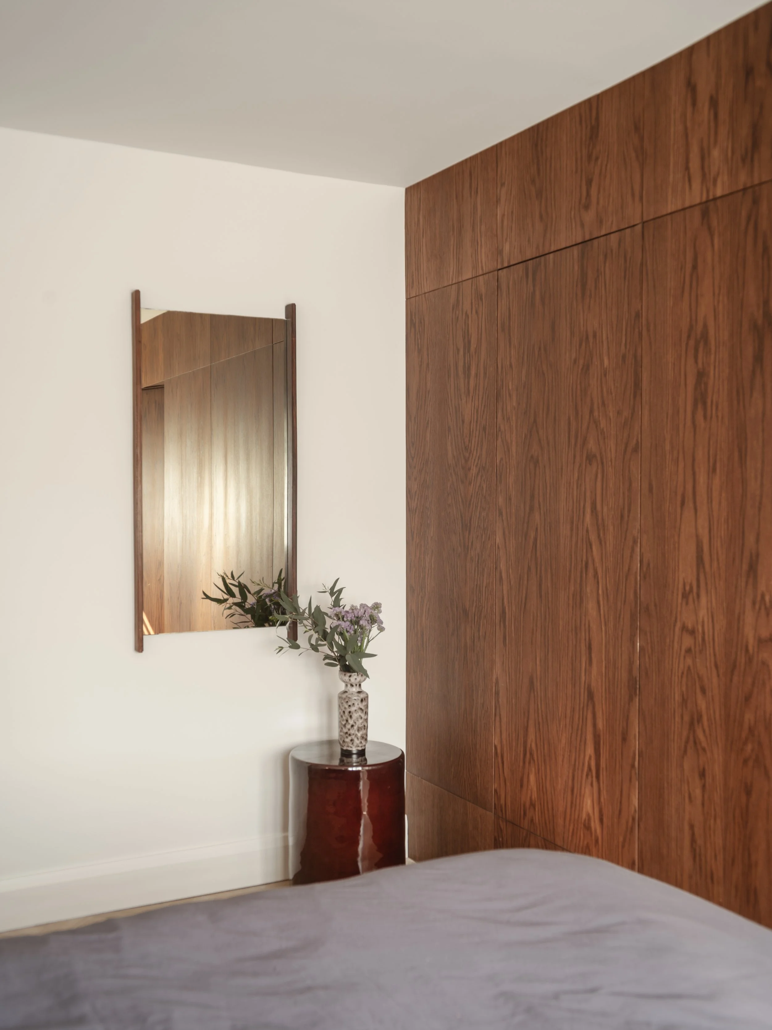 Bedroom with white wall, wooden headboard, mirror, vase with pink flowers on a dark red side table.