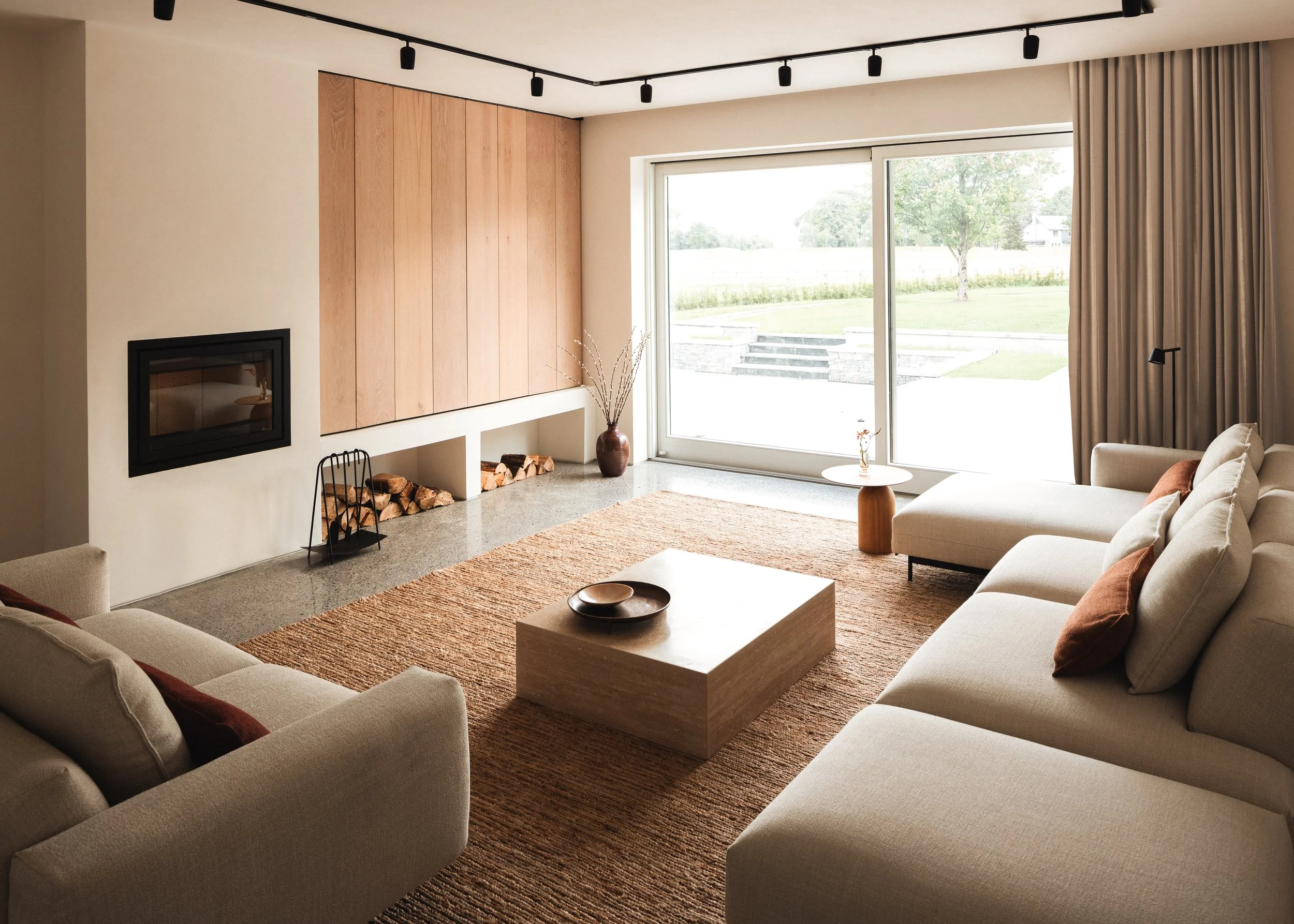 Modern living room with beige sectional sofa, wooden coffee table with bowls, large window with curtains, fireplace, and built-in wood storage, overlooking a green lawn.