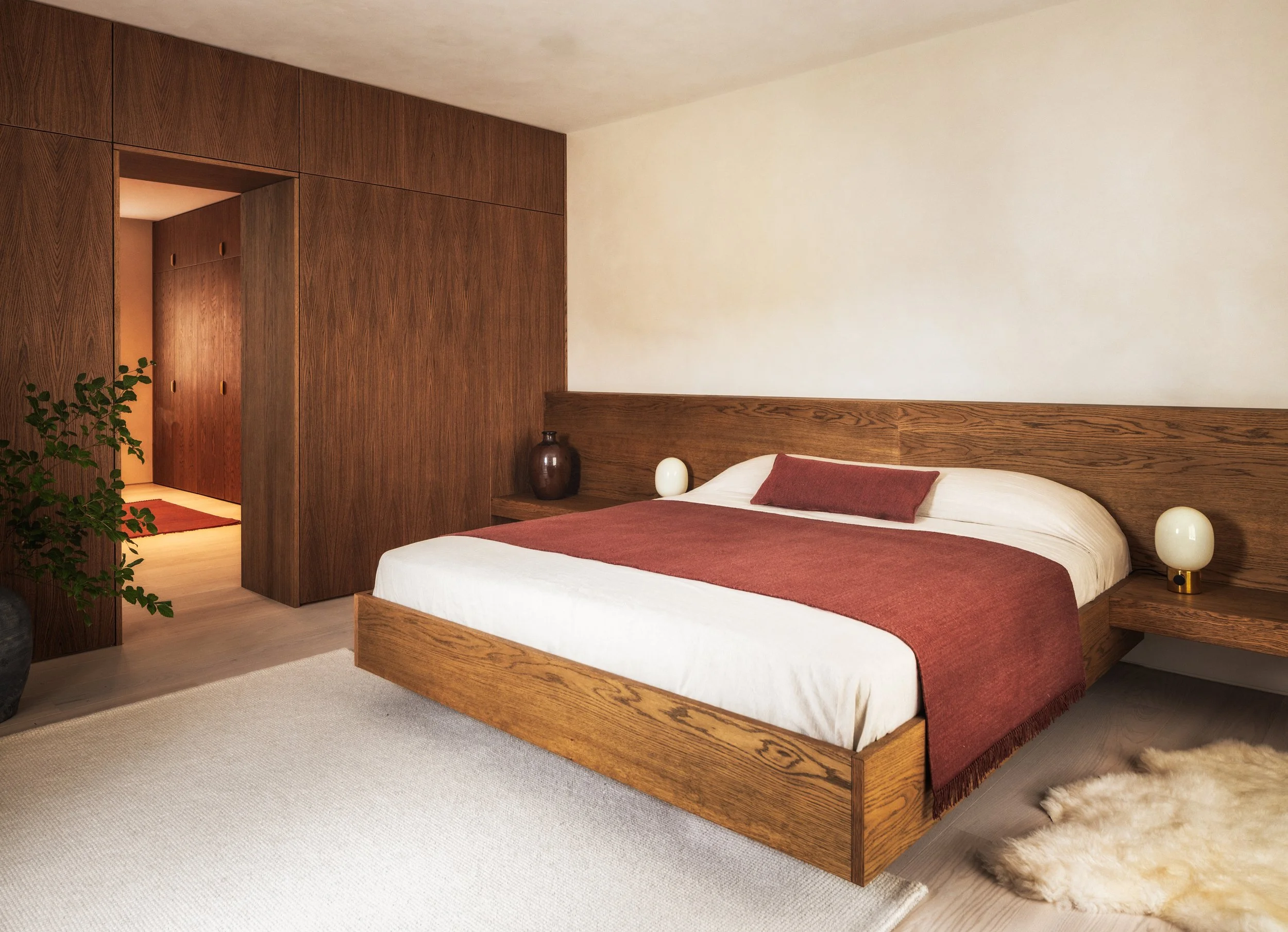 A bedroom with a wooden bed frame, a white bedspread, a rust-colored bed runner and pillow, two bedside lamps, a wooden wardrobe, and a plant in a pot near the door