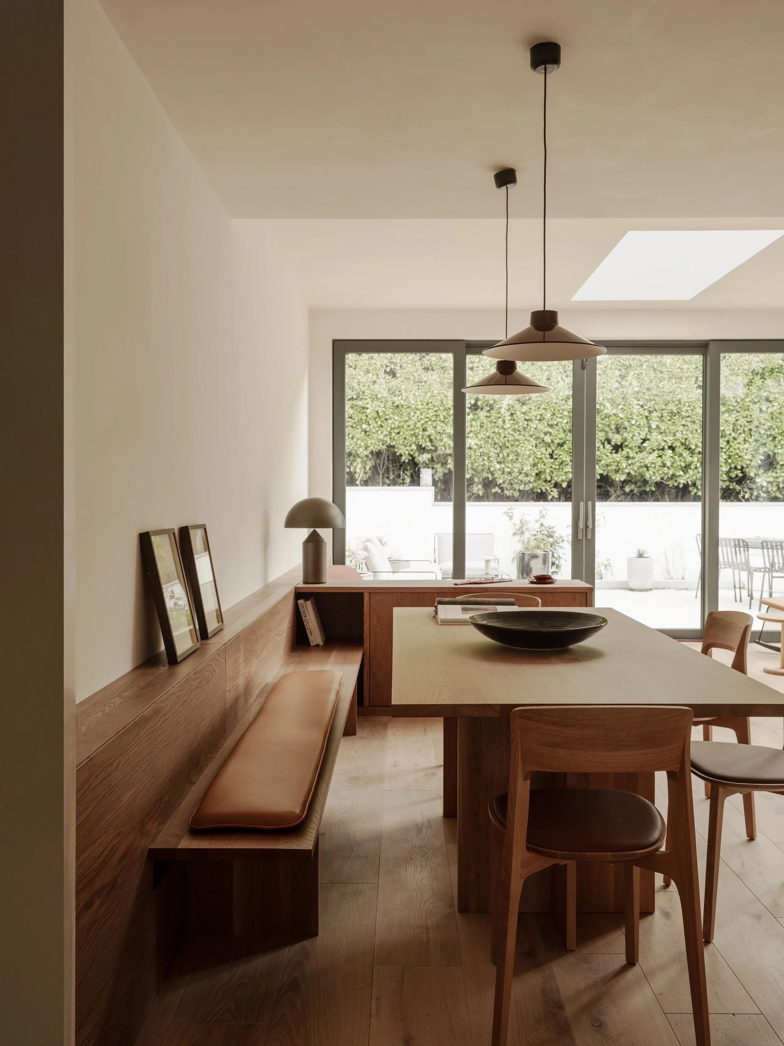 Modern dining room with wooden furniture, a bench with a leather cushion, and chairs around a wooden table. Large sliding glass doors lead to a patio with outdoor seating, and ceiling pendant lights hang over the table.