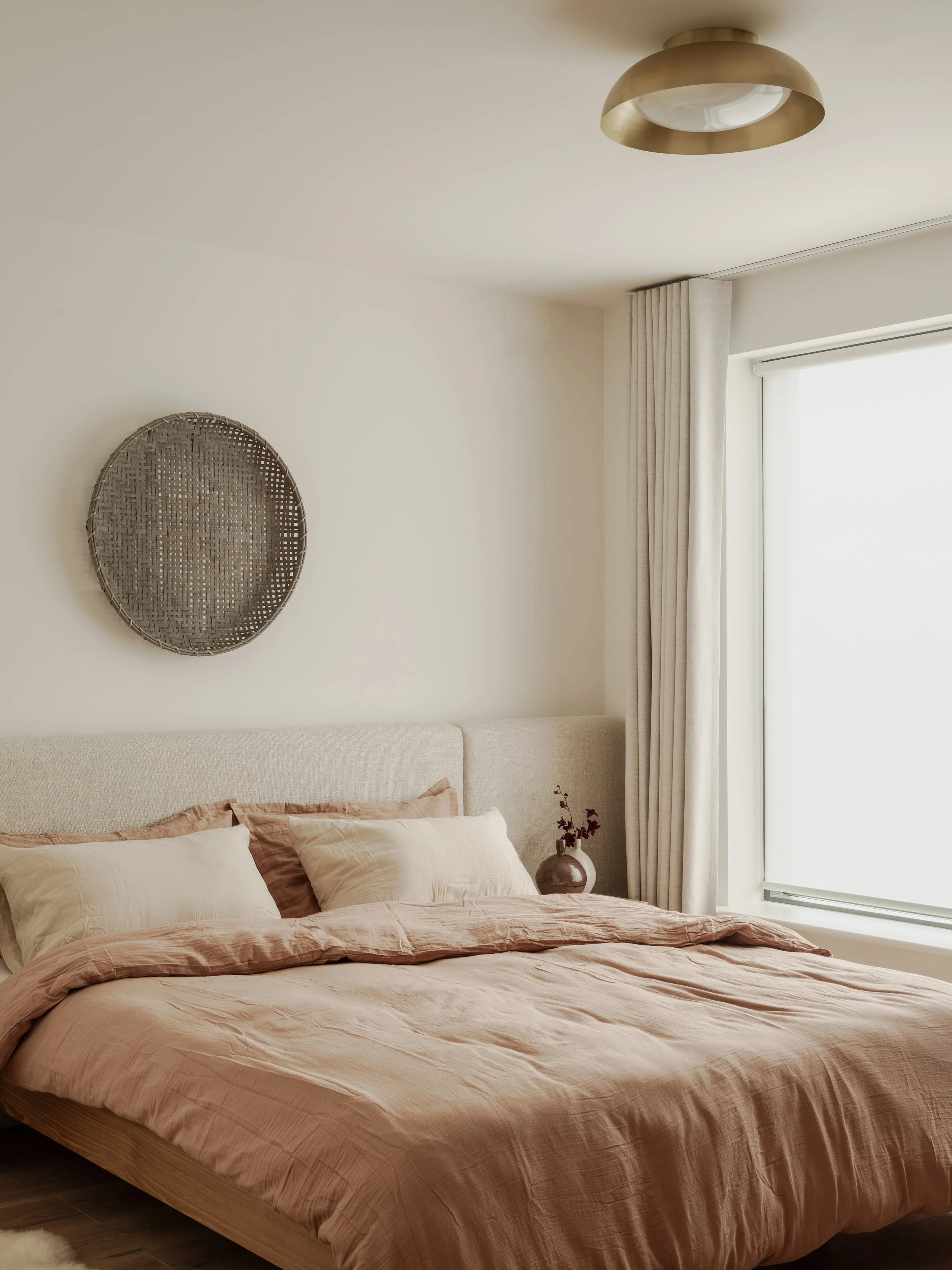 bedroom with dusty pink  and cream bed linen, bespoke fabric upholstered headboard with bamboo weaving hung above , brown and cream vases 
