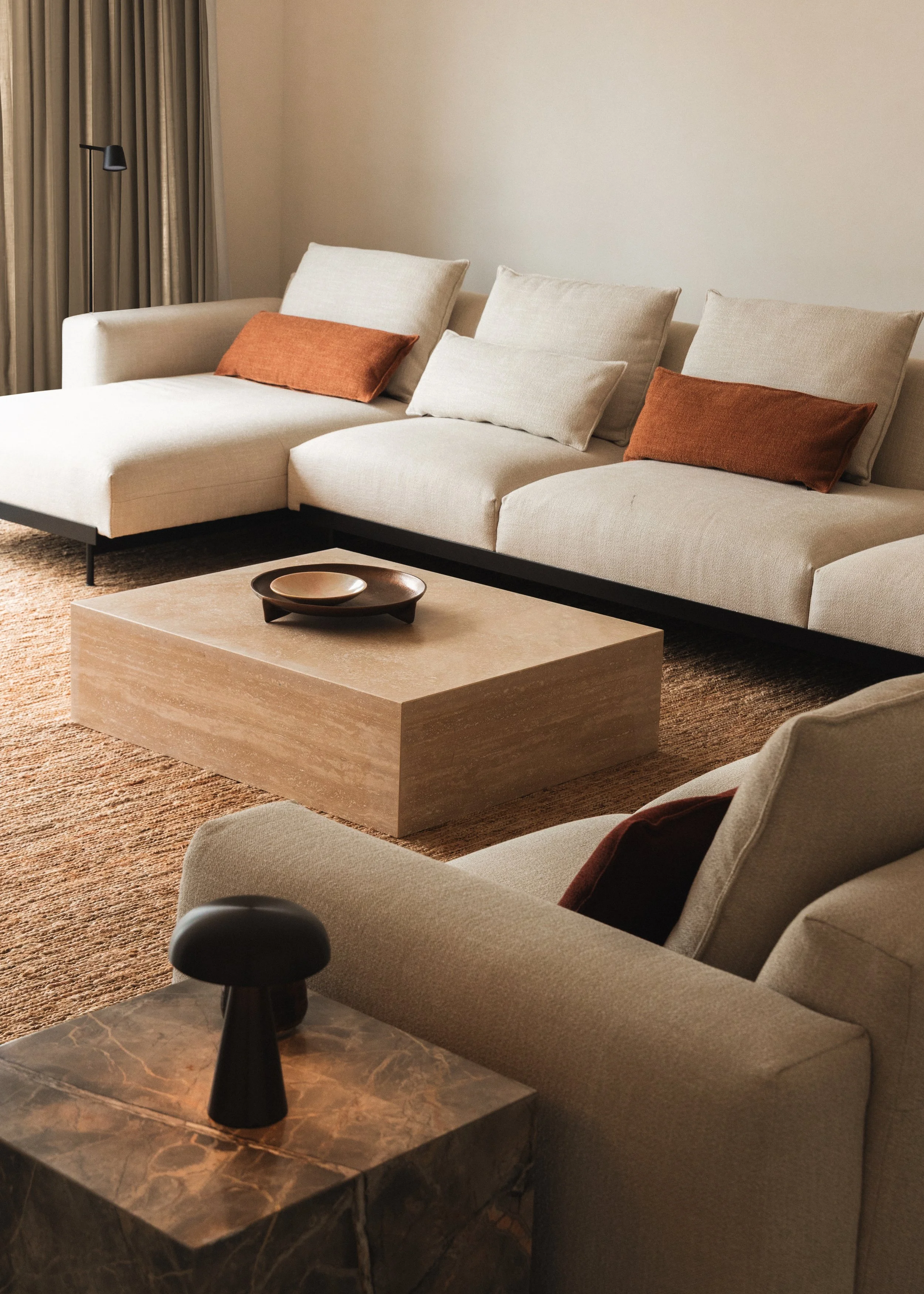 Interior of a living room with a cream-colored sectional sofa, brown pillows, a beige armchair, a rectangular stone coffee table, and a small black table lamp on a marble side table.