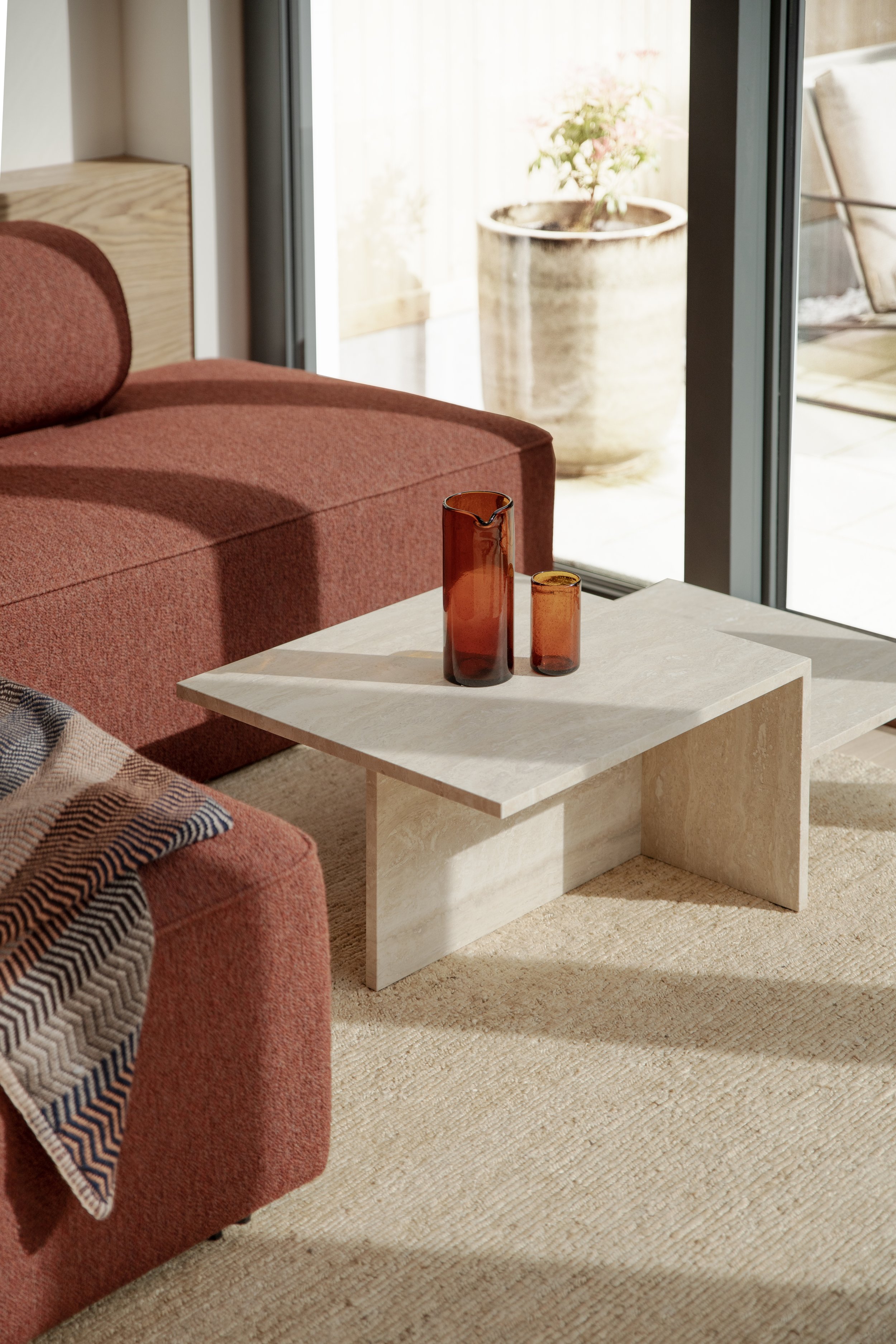 A living room with a red sofa, a beige coffee table with two amber-colored vases, a window with a sliding door, and a large potted plant outside.