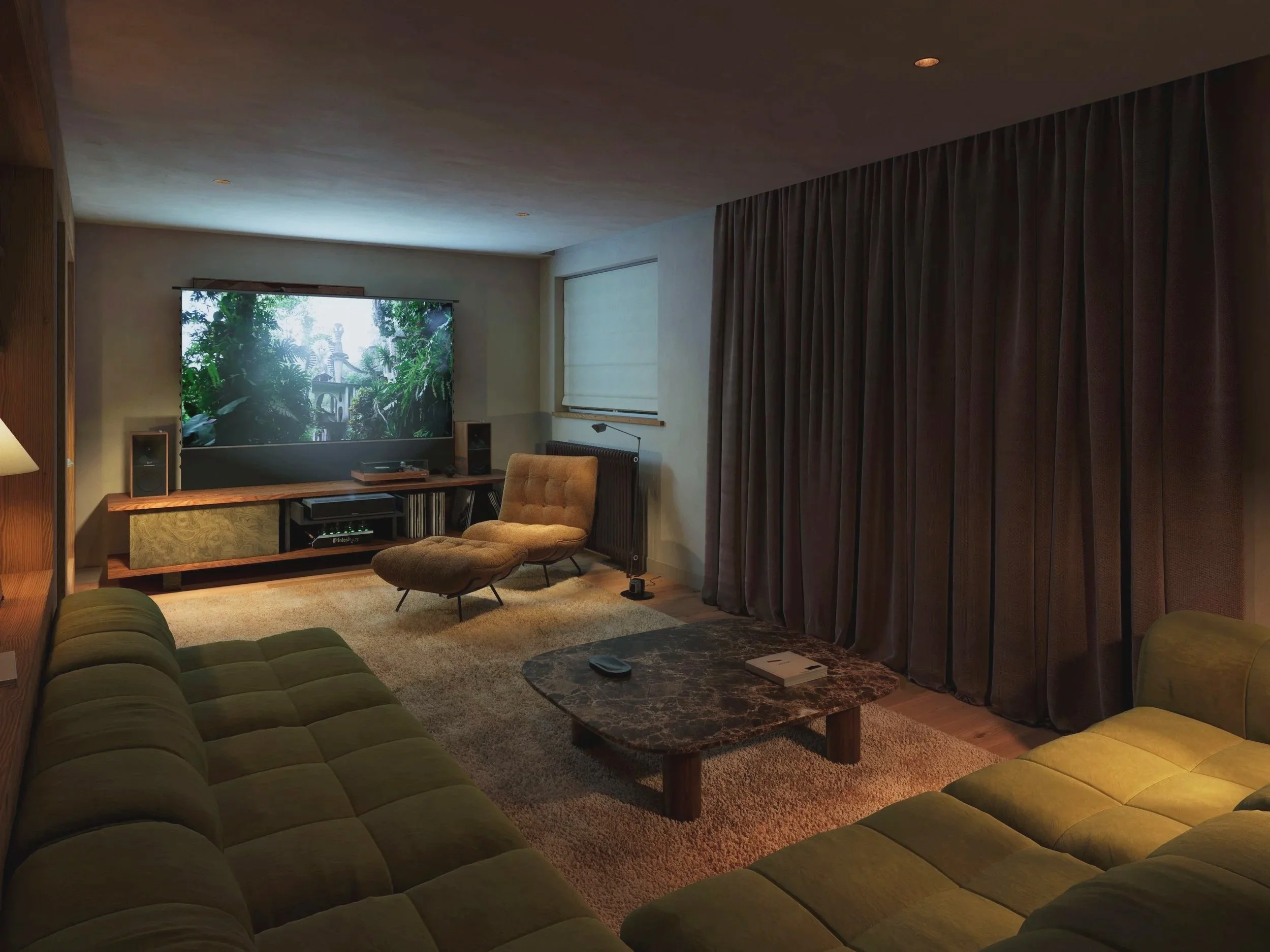 Living room with olive-green sectional sofa, beige armchair with ottoman, marble coffee table, large flat-screen TV, and dark heavy curtains covering the window.