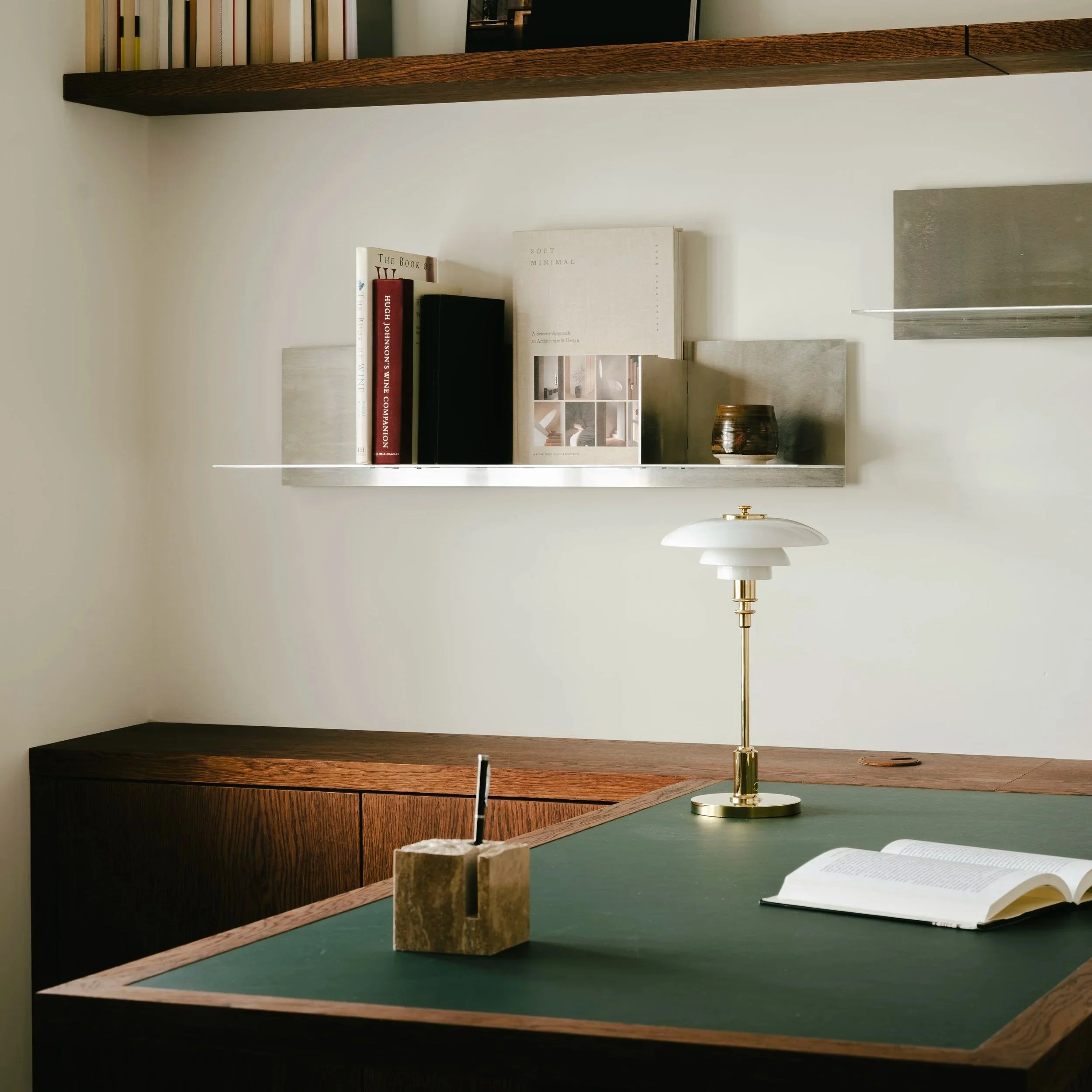 A desk with a green surface, a white and gold desk lamp, an open book, and a stone pencil holder with a pen. Above the desk, a floating shelf with books and a small decorative container, set against a plain wall.