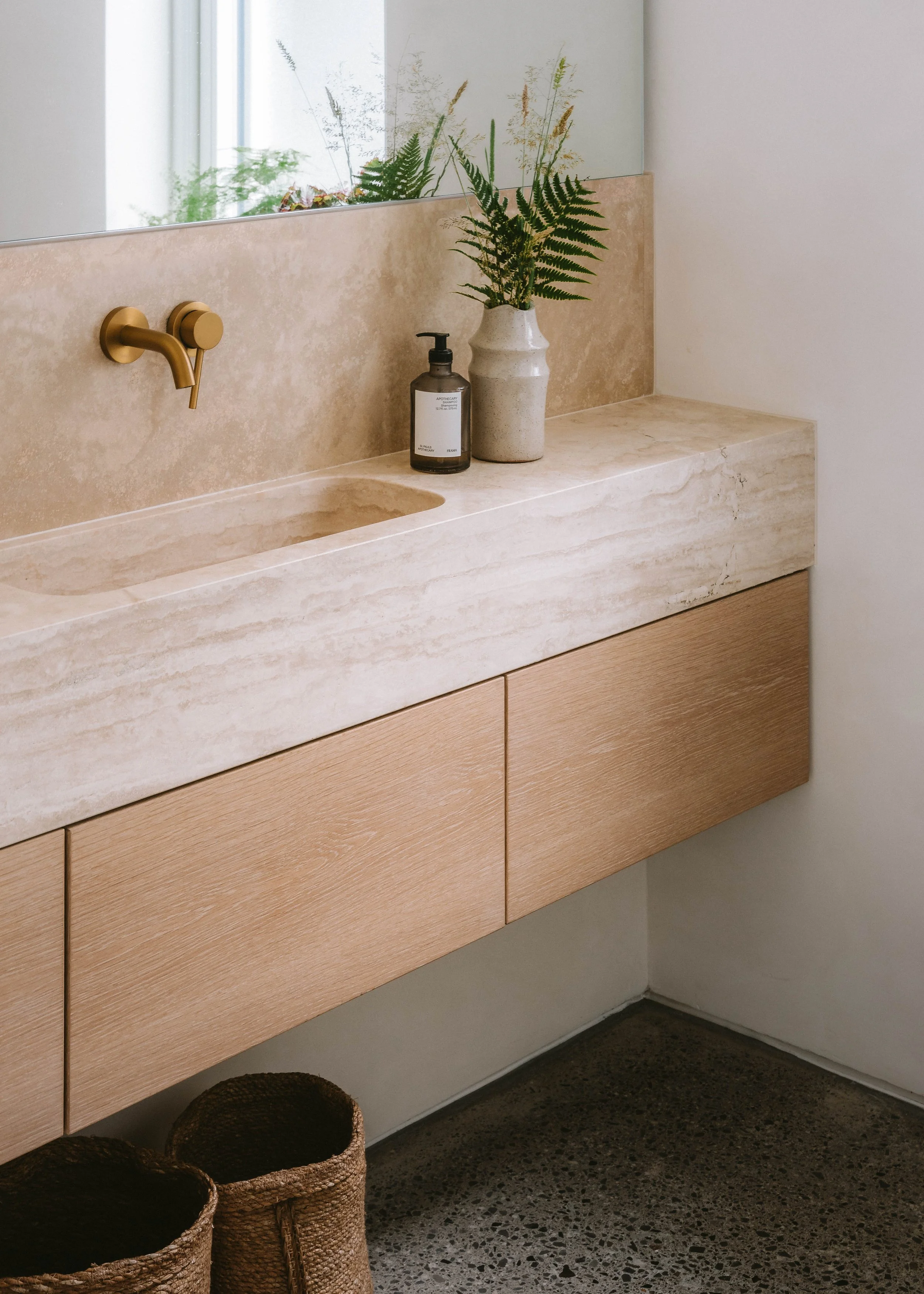 Bathroom vanity with a beige stone countertop, wooden cabinet, a black soap dispenser, a vase with green fern leaves, a mirror, and two woven baskets on a speckled concrete floor.