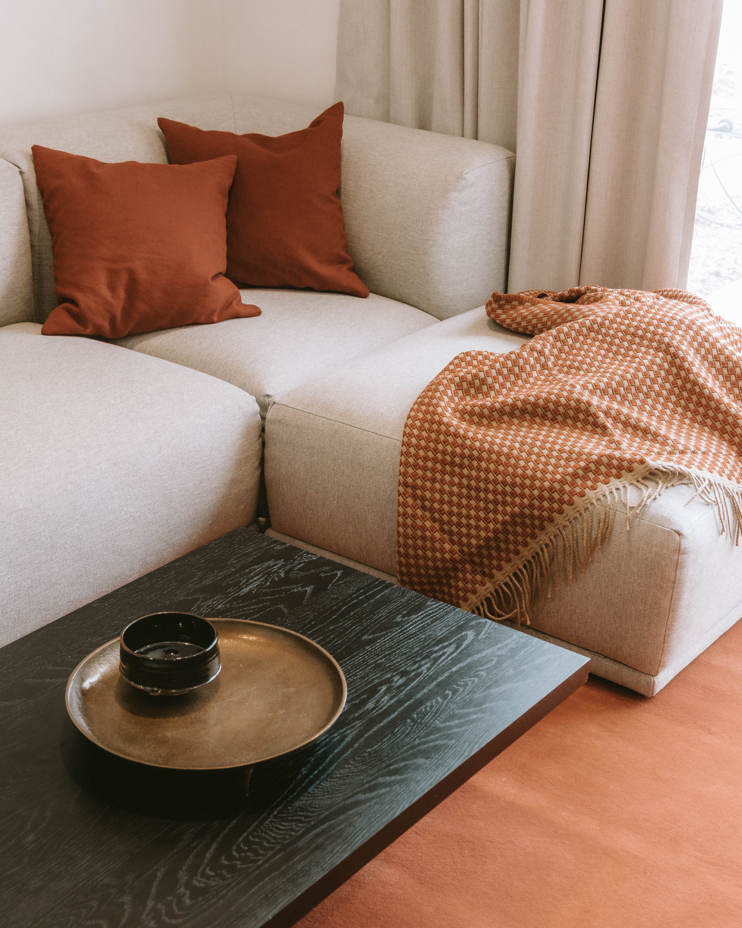 A cozy living room corner with a beige sectional sofa, two rust-colored throw pillows, an orange patterned blanket draped over the sofa, a black coffee table with a bronze plate and black bowl, beige curtains, and a light-colored wall.