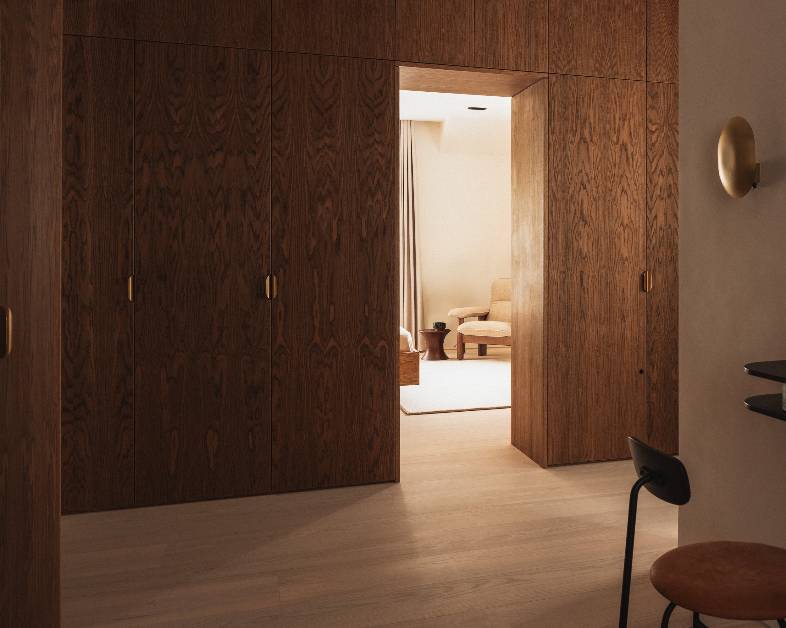Interior view of a room with wooden walls and an open doorway leading to a lounge area with beige chairs, a small table, and floor-to-ceiling curtains.