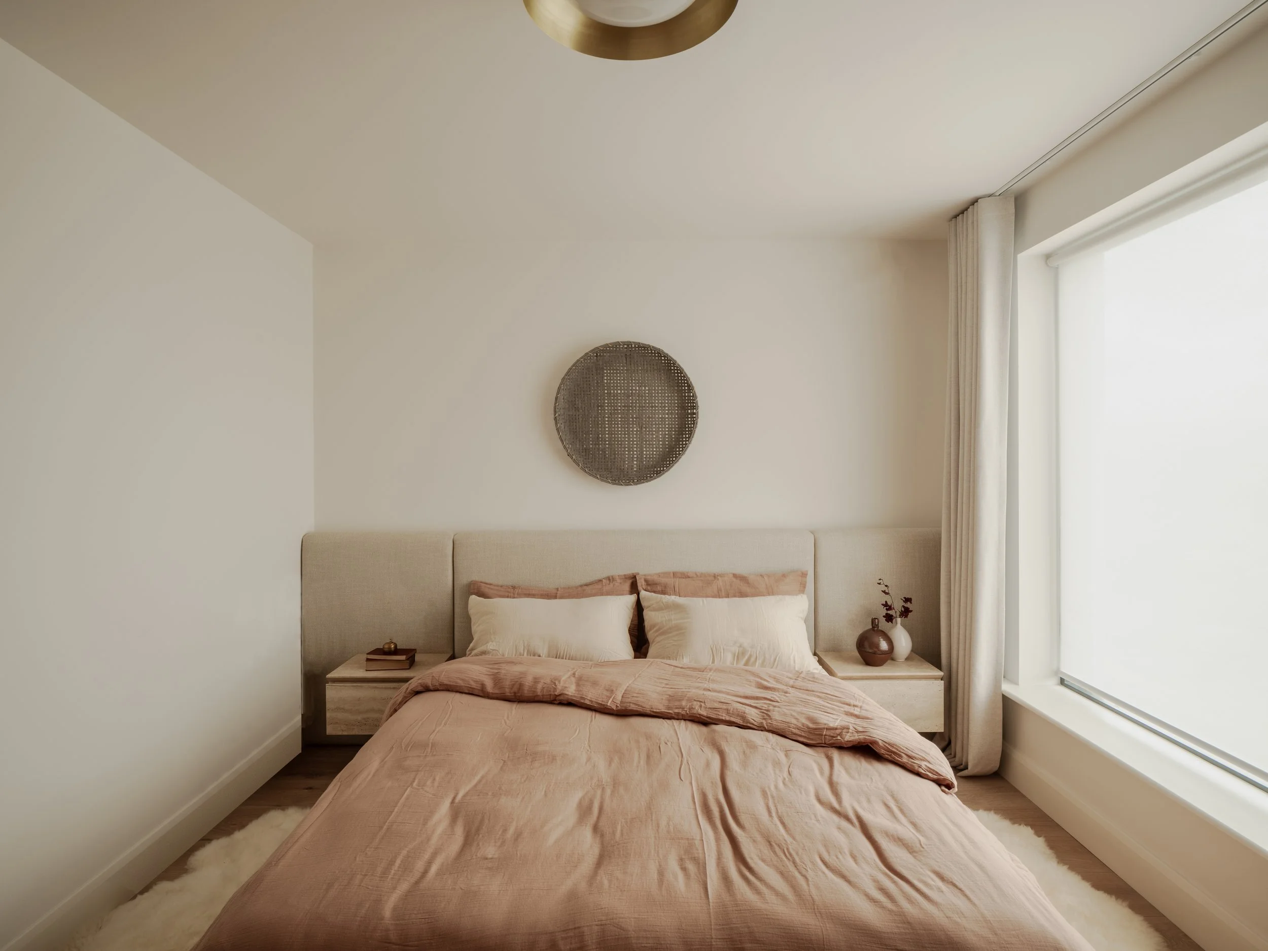 bedroom with dusty pink  and cream bed linen, bespoke travertine bedside tables,  bespoke fabric upholstered headboard with bamboo weaving hung above , brown and cream vases 