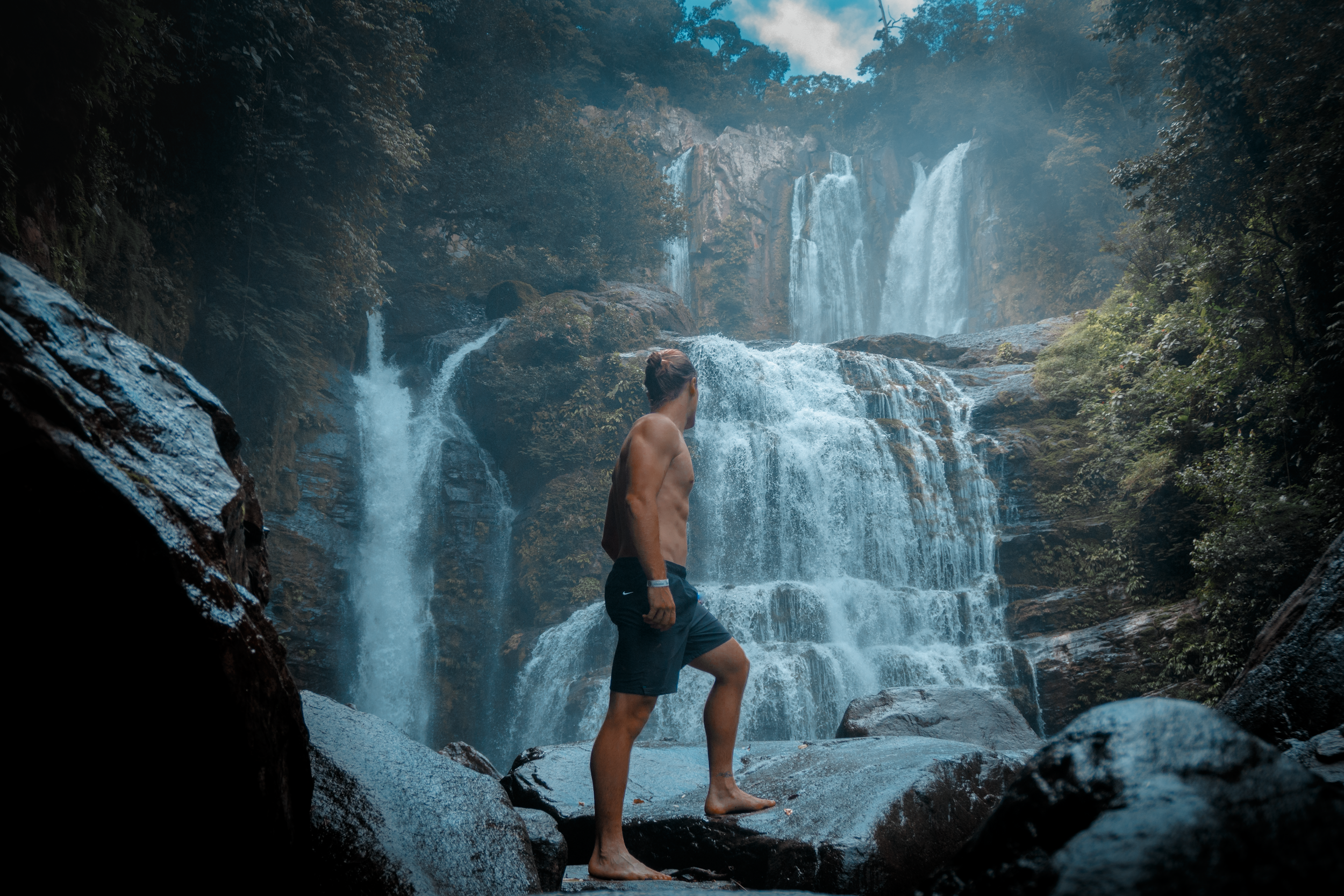 A shirtless man in shorts stands on a wet rock in front of a large cascading waterfall in a lush forest.