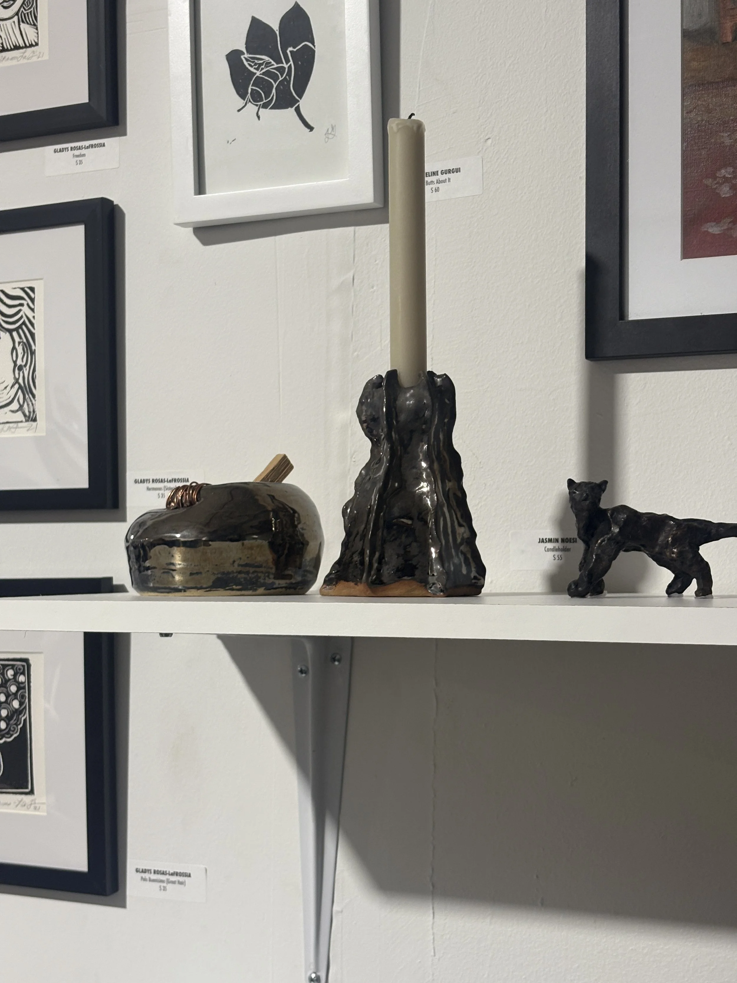 Display shelf with three black ceramic sculptures: one in the shape of an apple, one in the shape of a tall candle holder, and one in the shape of a dog. Framed artwork is on the wall behind the shelf.