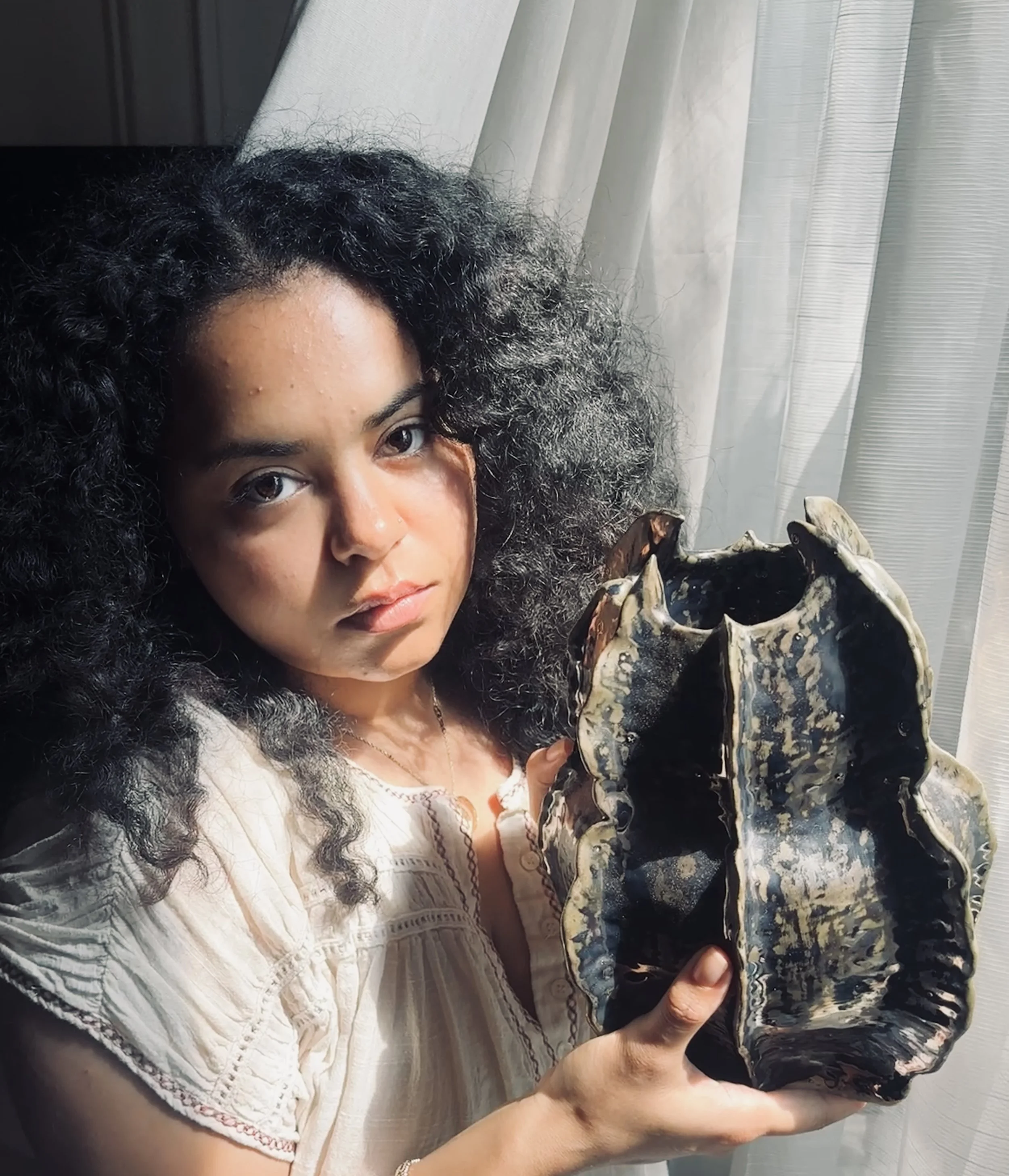 A woman with curly black hair holding a turtle with a patterned shell, near a window with sheer curtains.