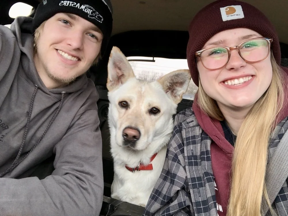 A smiling young man and woman with a white dog sitting between them in a vehicle.
