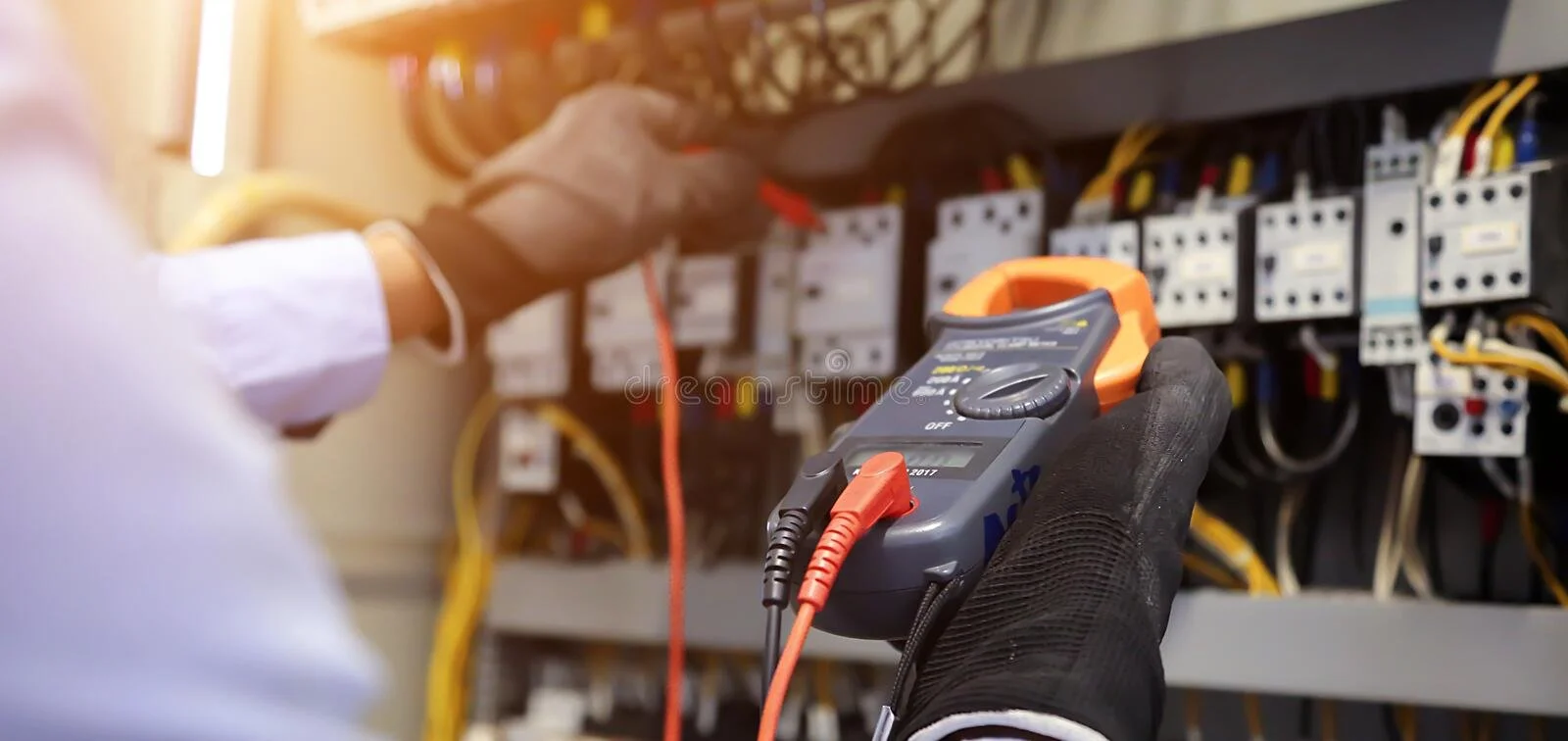 A technician wearing gloves is using a digital multimeter to test electrical wires inside an electrical panel.