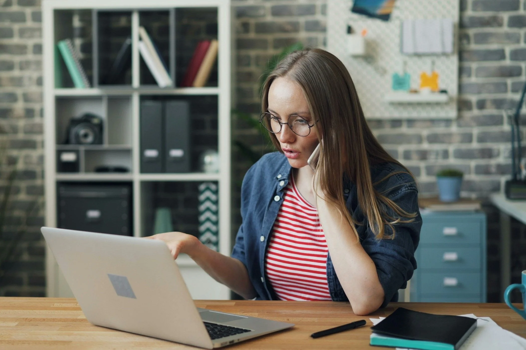Woman with glasses sitting at a desk in an office, talking on her phone, with a laptop in front of her, and bookshelf in the background.