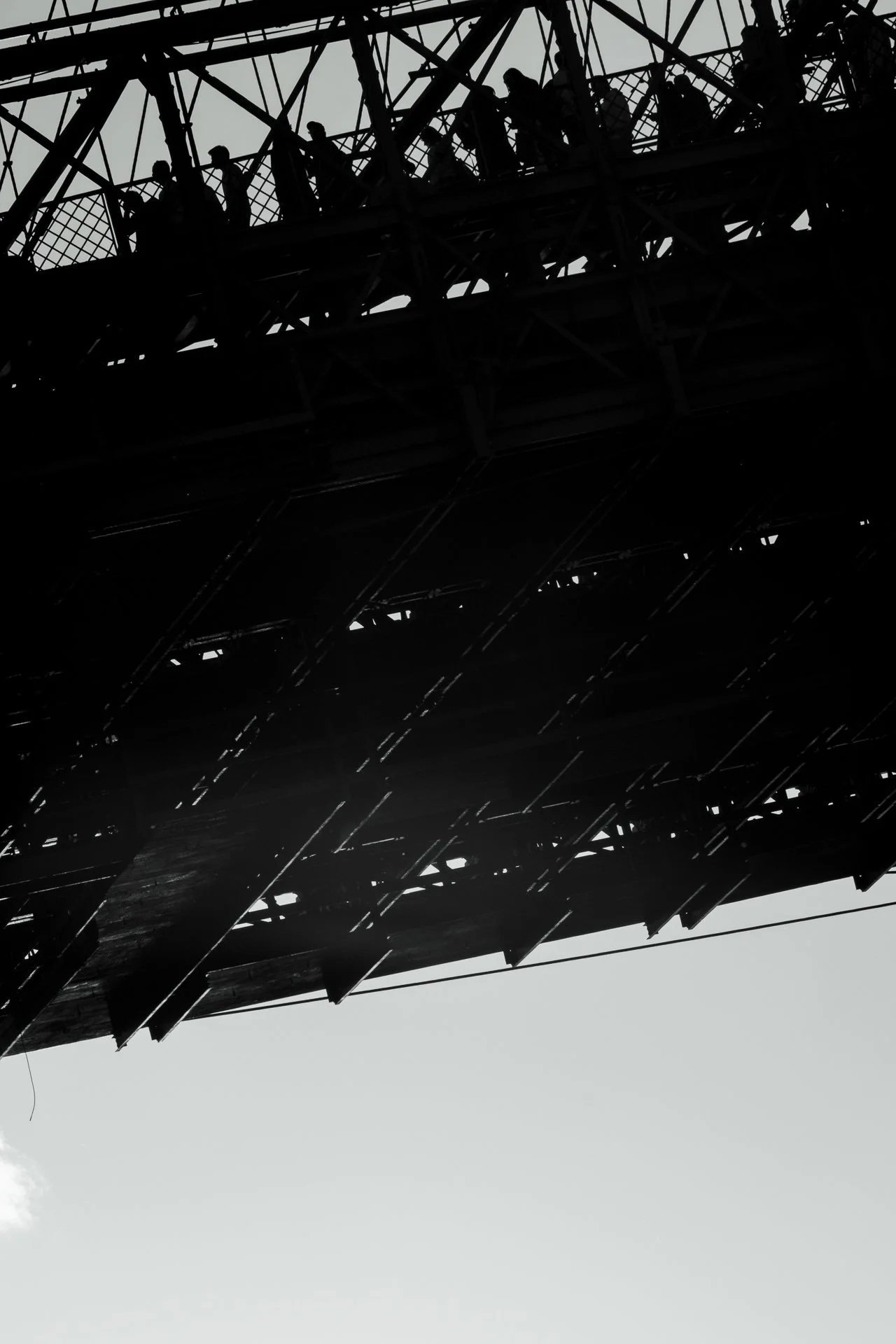 Silhouette of people walking on a bridge or platform with a complex metal lattice structure against a light sky.