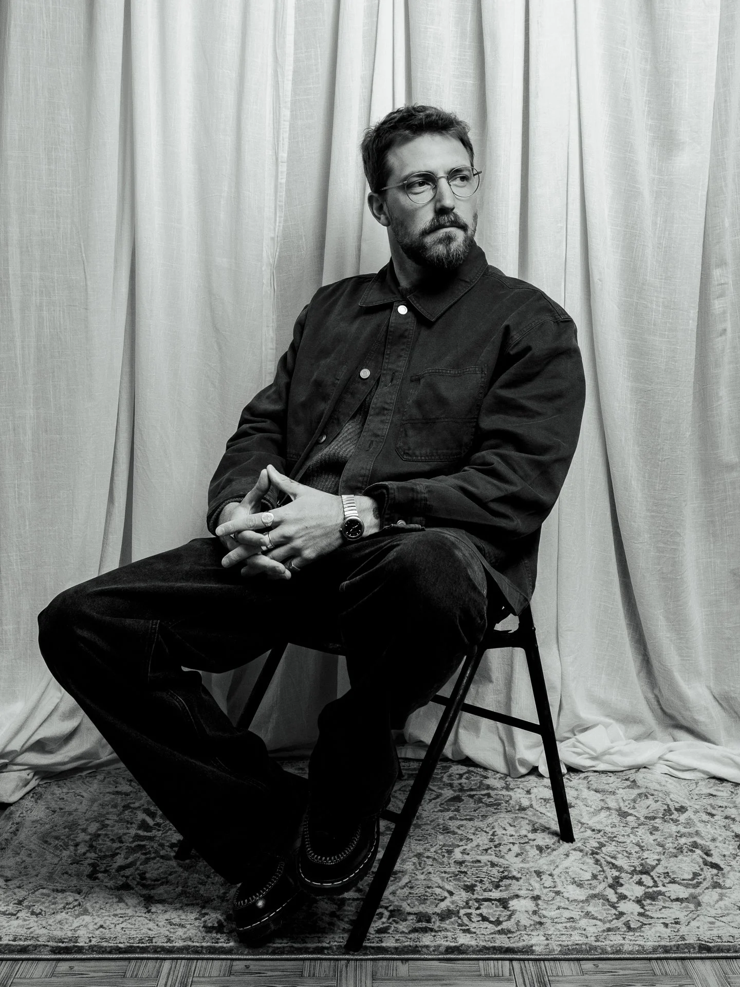 black and white portrait photography natural light studio of a man sitting on a chair in front of a curtain. He is wearing glasses, a jacket, and has a beard. He is looking to his right with his hands clasped.
