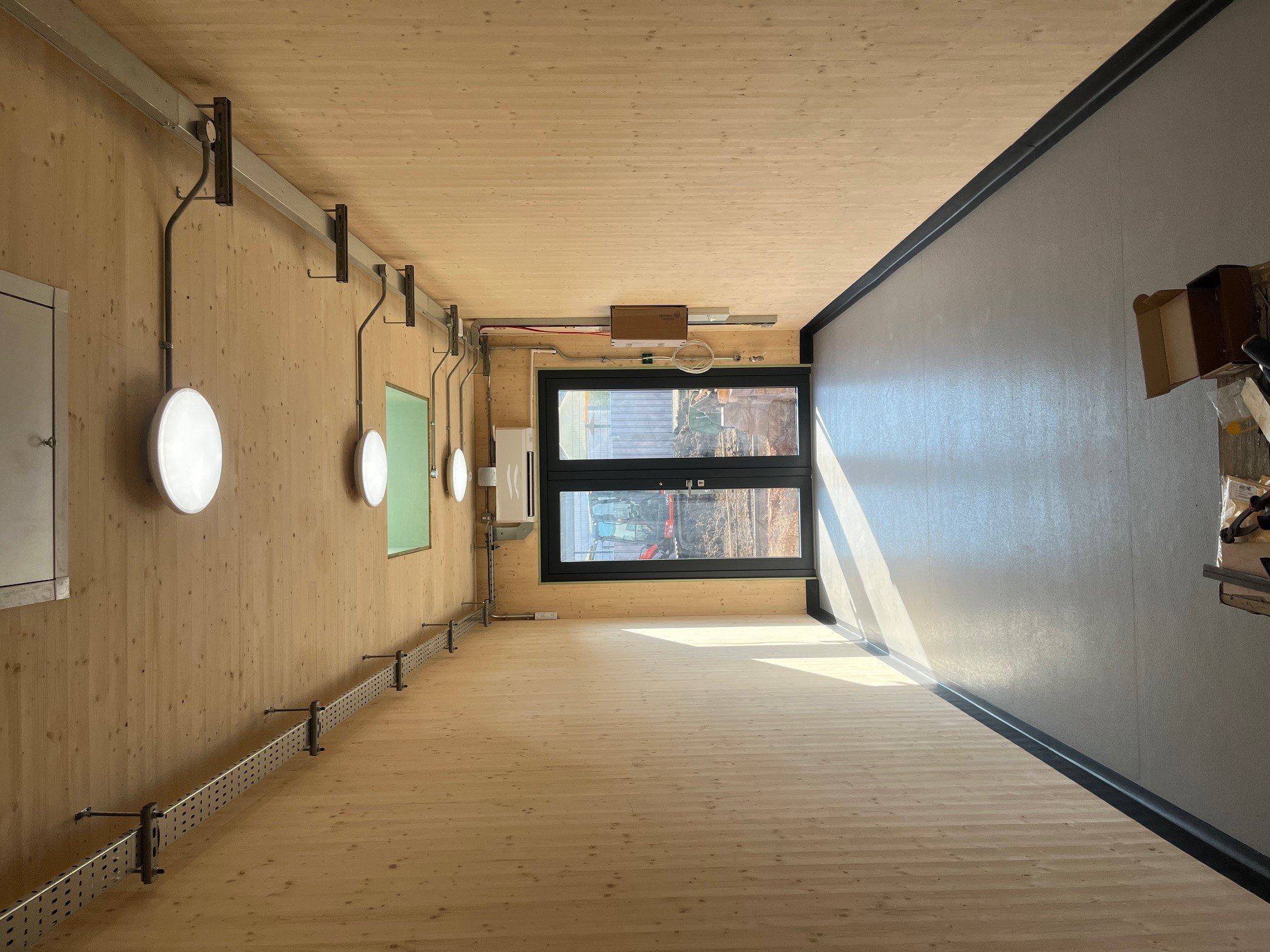 Empty room with wooden walls and floor, large glass door, and some technical equipment or construction features along one wall.