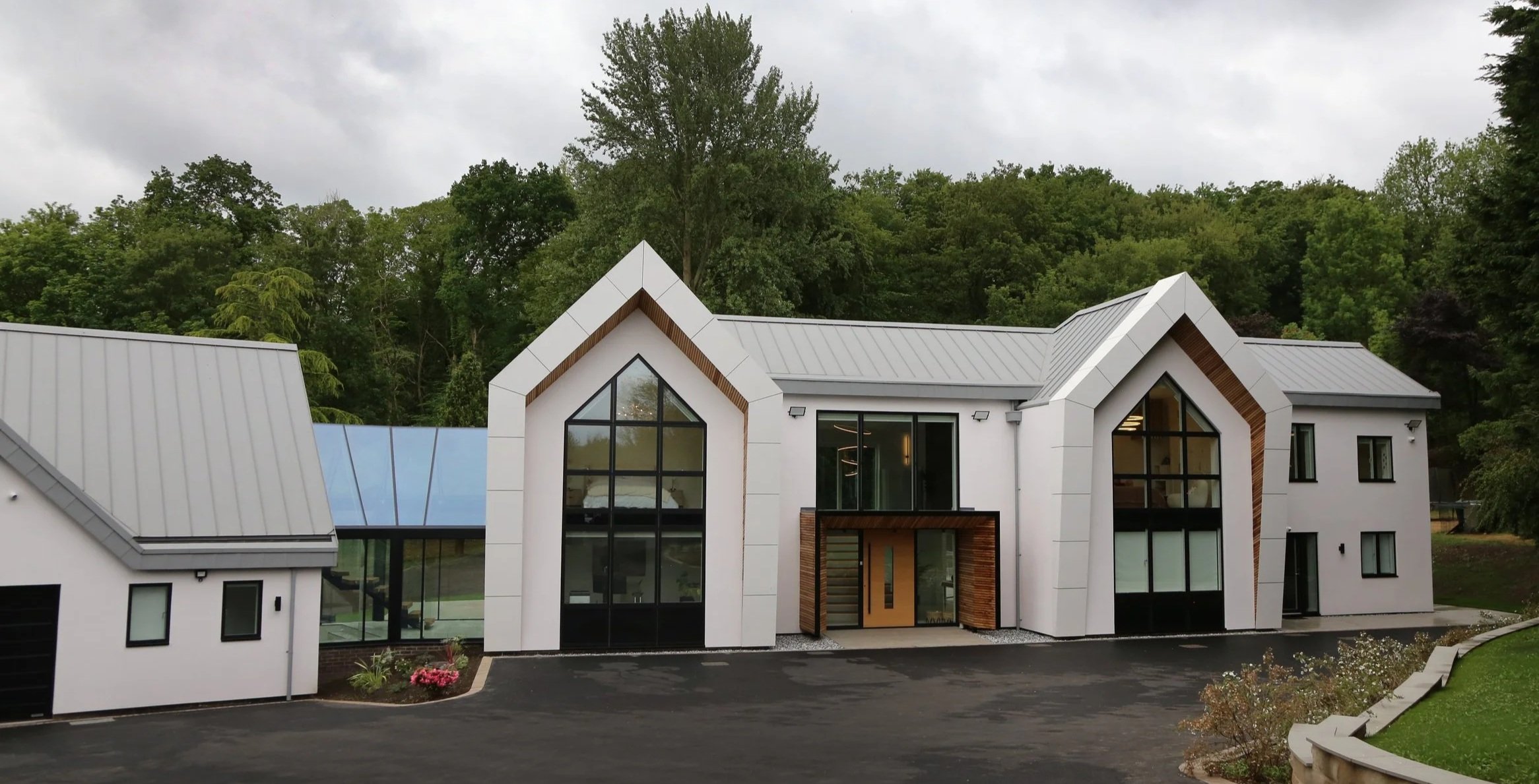 A modern two-story house with large windows, white exterior walls, and gray metal roofs, situated in a wooded area with a paved driveway in front.