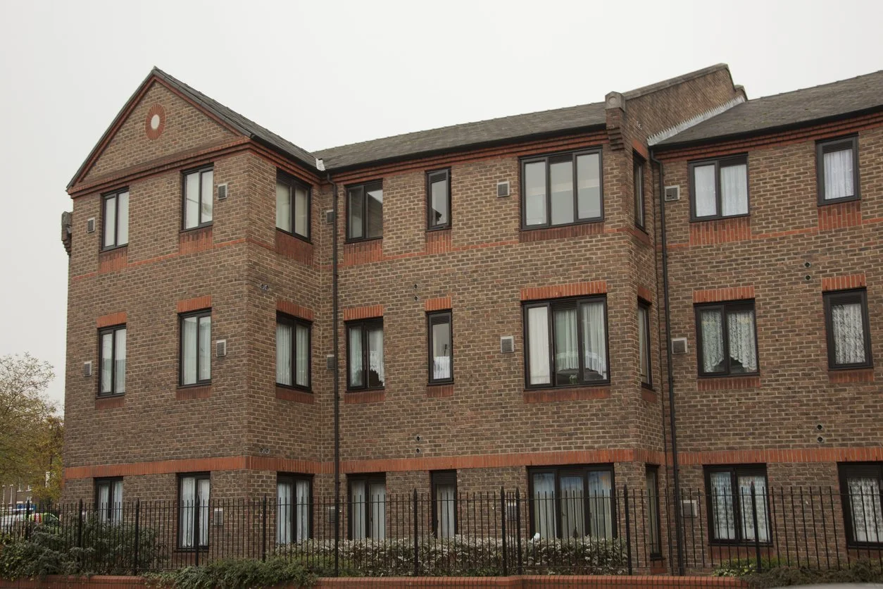 A brick apartment building with multiple windows and a black metal fence in the foreground.