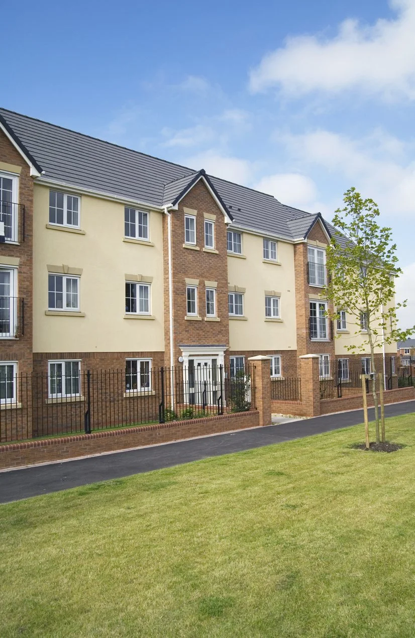 A multi-story apartment building with brick and beige exterior walls, white-framed windows, and a black metal fence in the front yard. There is a small tree and a paved walkway along the building with a grassy lawn in the foreground. The sky is partly cloudy with blue sky visible.