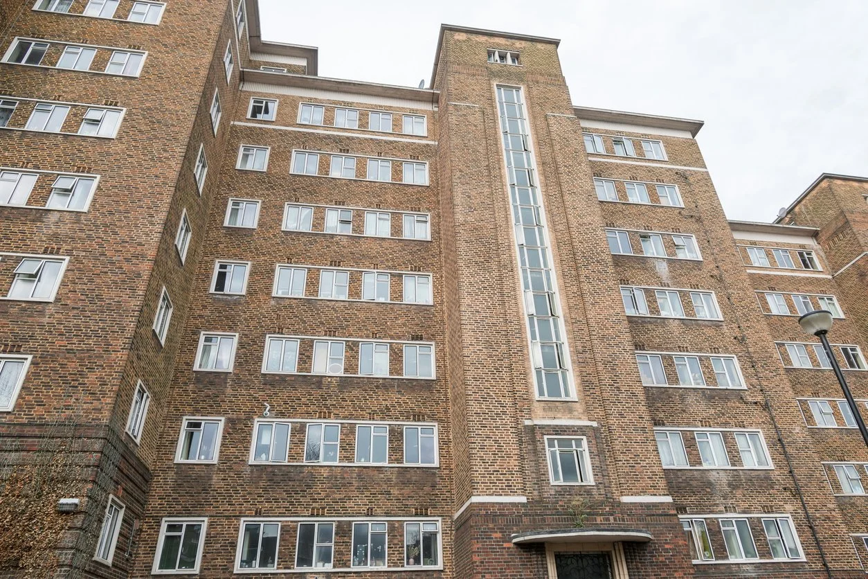 Low-angle view of a multi-story brick residential apartment building with many windows, a vertical strip of glass windows in the center, and a building entrance at the bottom.