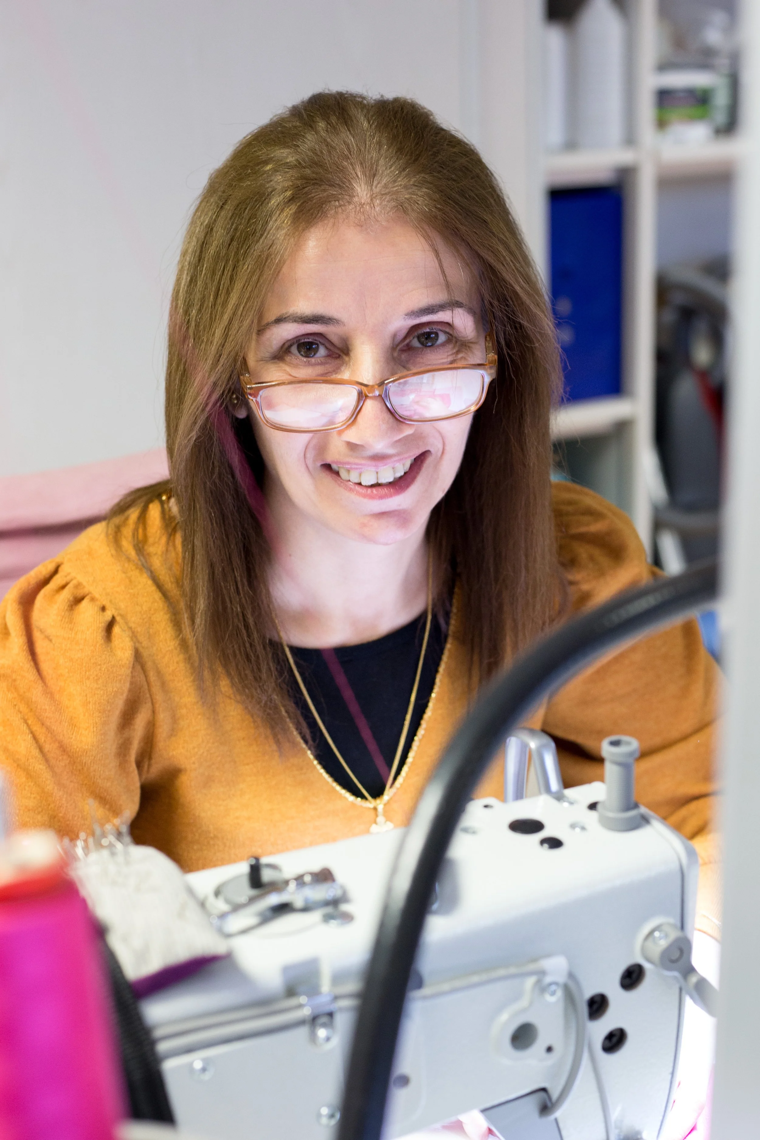 Femme souriante portant des lunettes, travaillant à une machine à coudre dans un atelier.