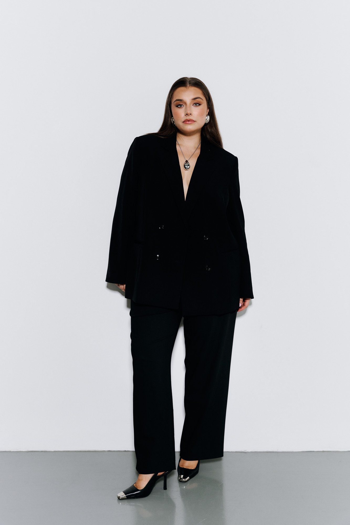 A woman with long brown hair in a black pantsuit with a deep V-neck shirt, wearing a necklace and earrings, standing against a plain white background.