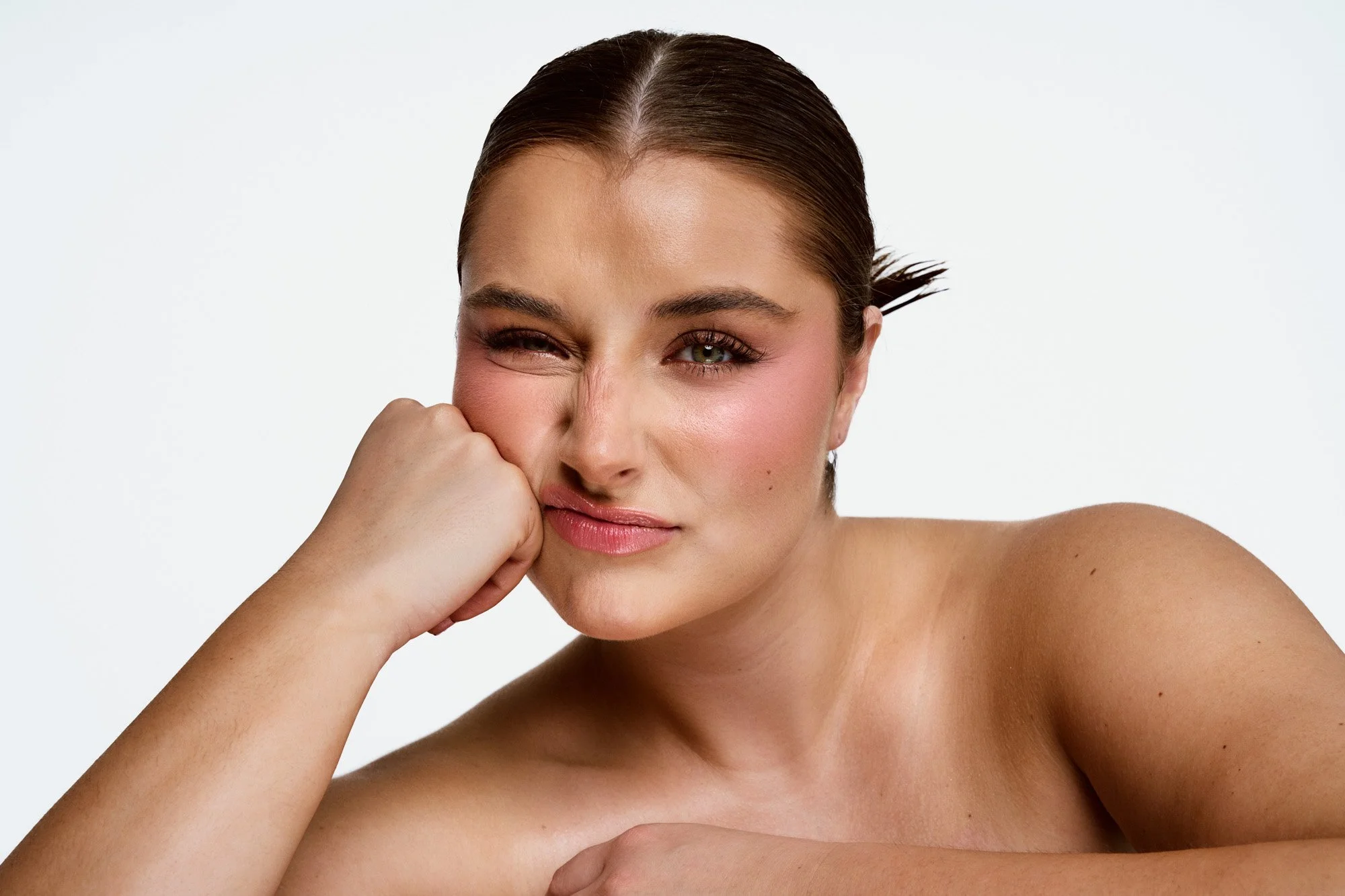 Close-up of a woman with wet brown hair, playful expression, one eye winking, resting her chin on her hand, skin makeup with blush and glossy lips, against a plain white background.