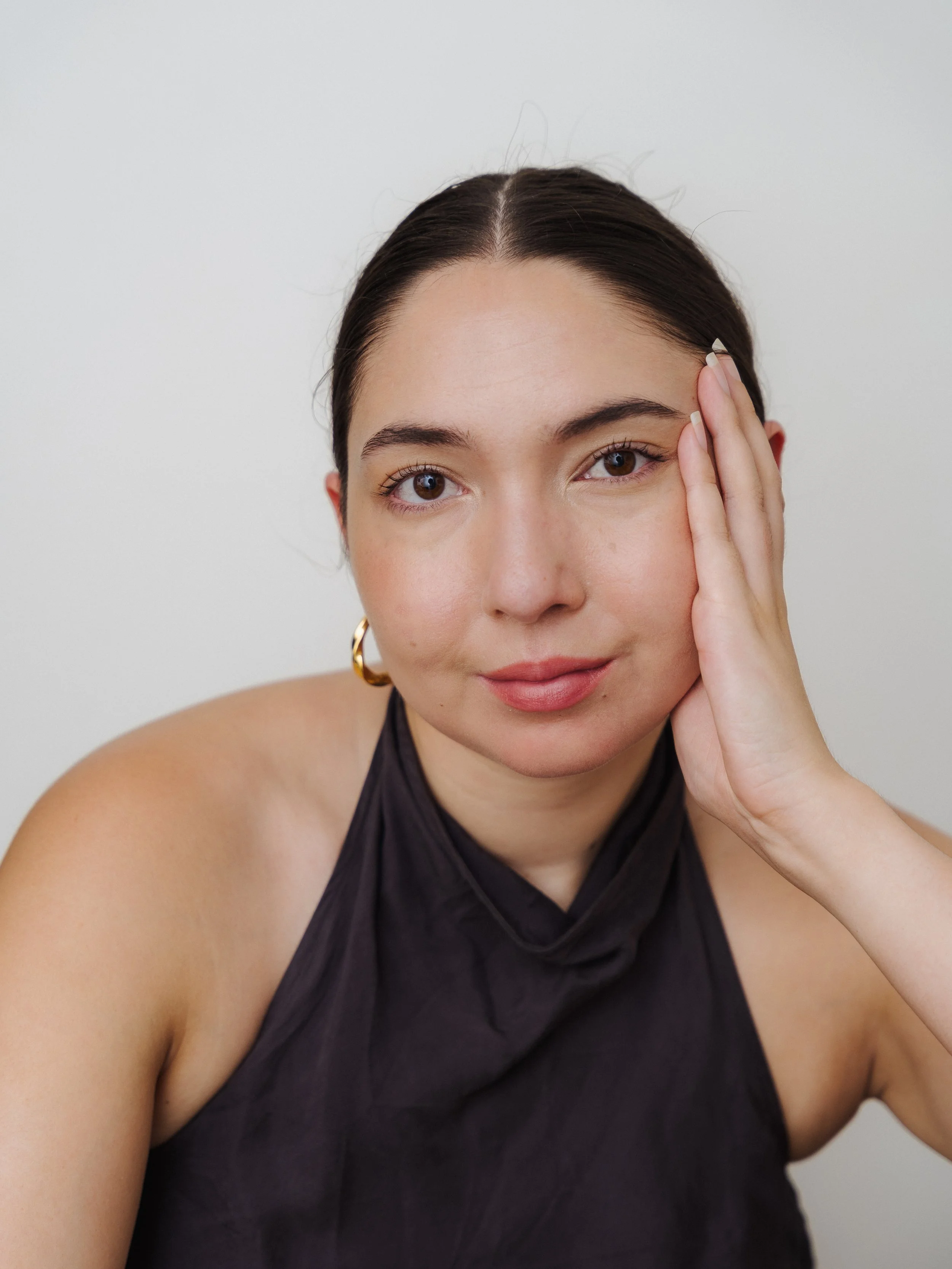 A woman, Wilhelmina Martens, with dark hair pulled back, wearing a black sleeveless top and gold hoop earrings, resting her face on her left hand against a plain white background.